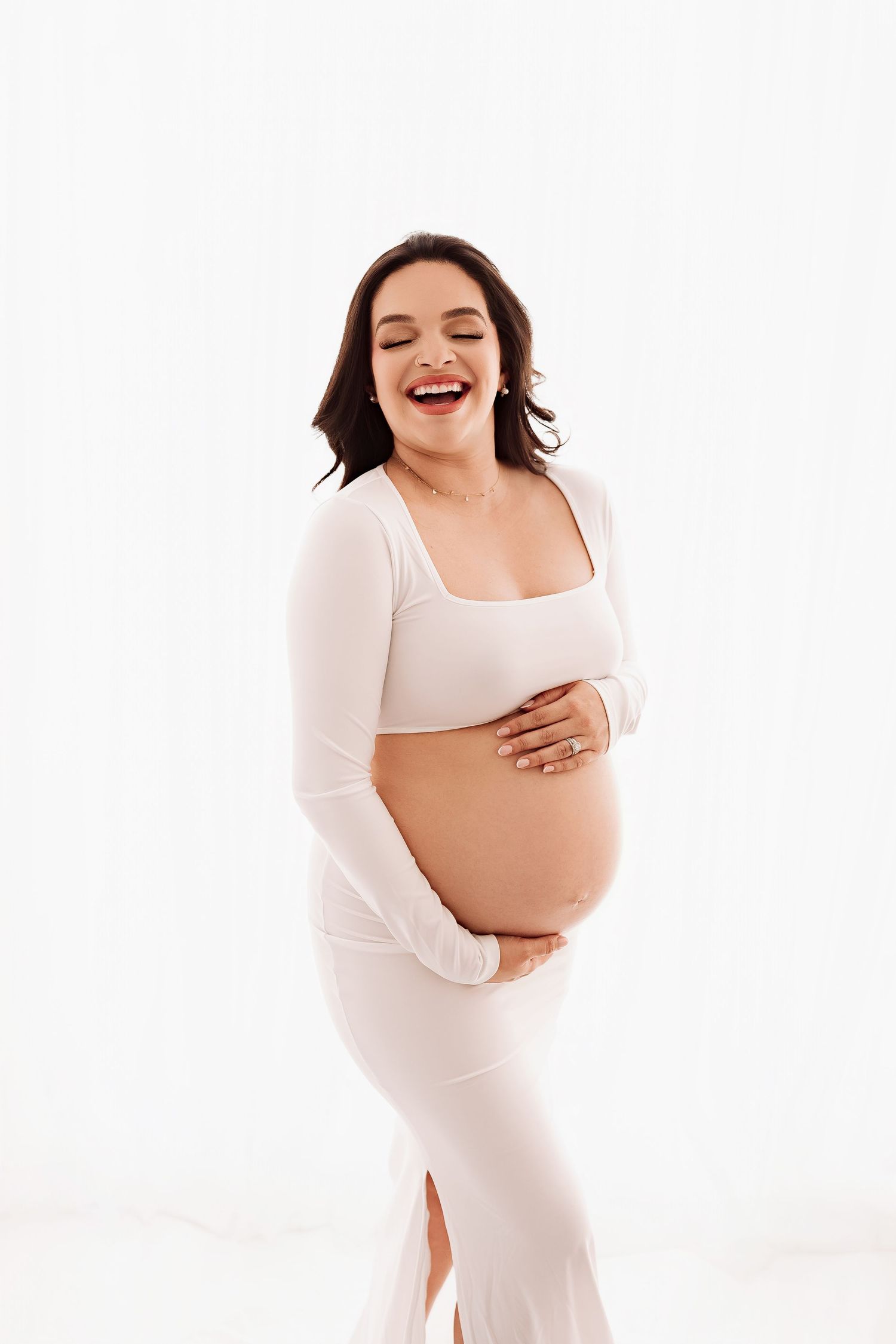 Joyful pregnant woman in white dress laughing while cradling her bare baby bump.