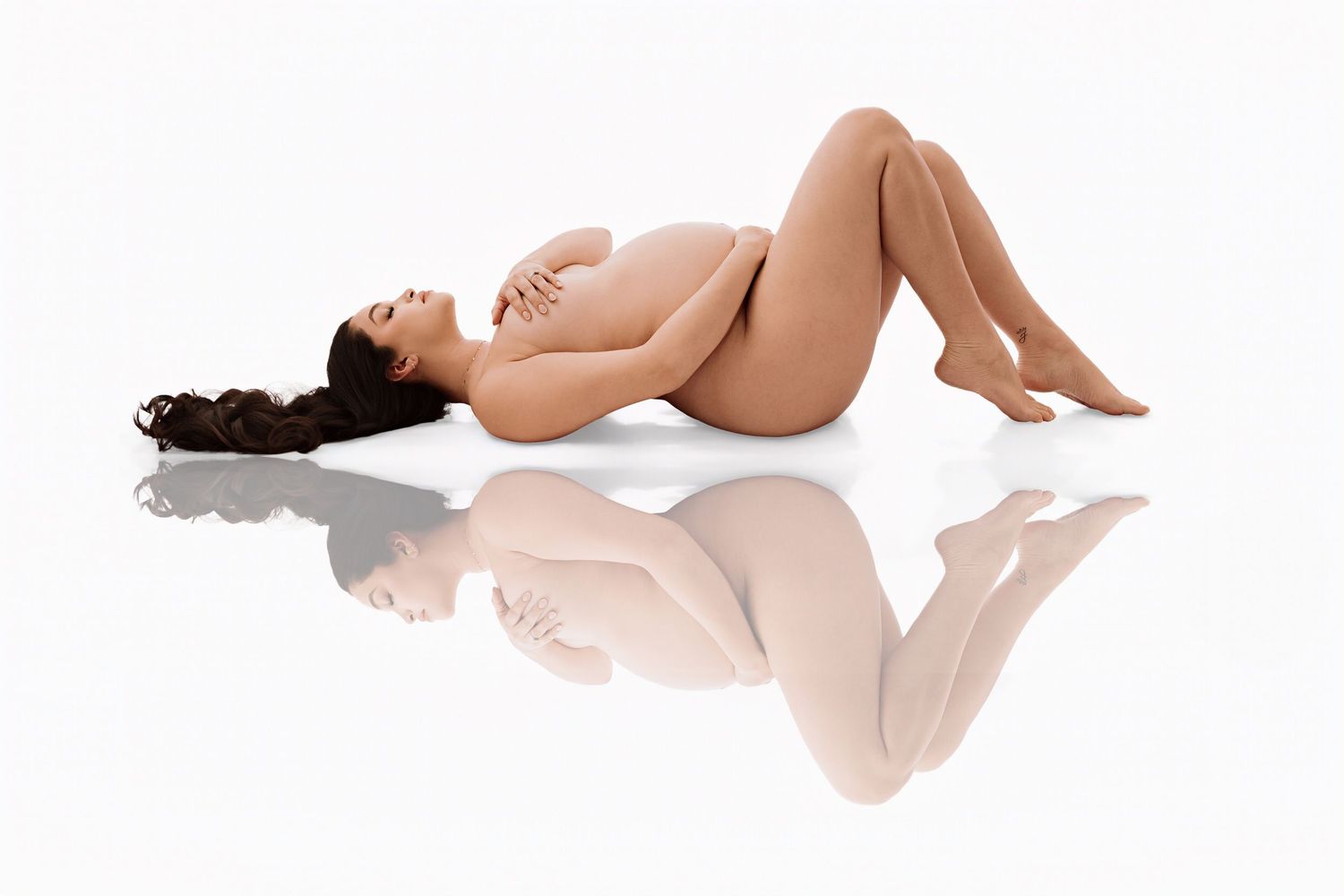 Nude pregnant woman lying on reflective white surface, cradling baby bump in artistic maternity photo.