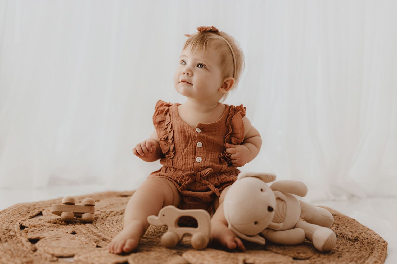Curious baby girl in rust romper looking up while sitting on woven rug with toys.