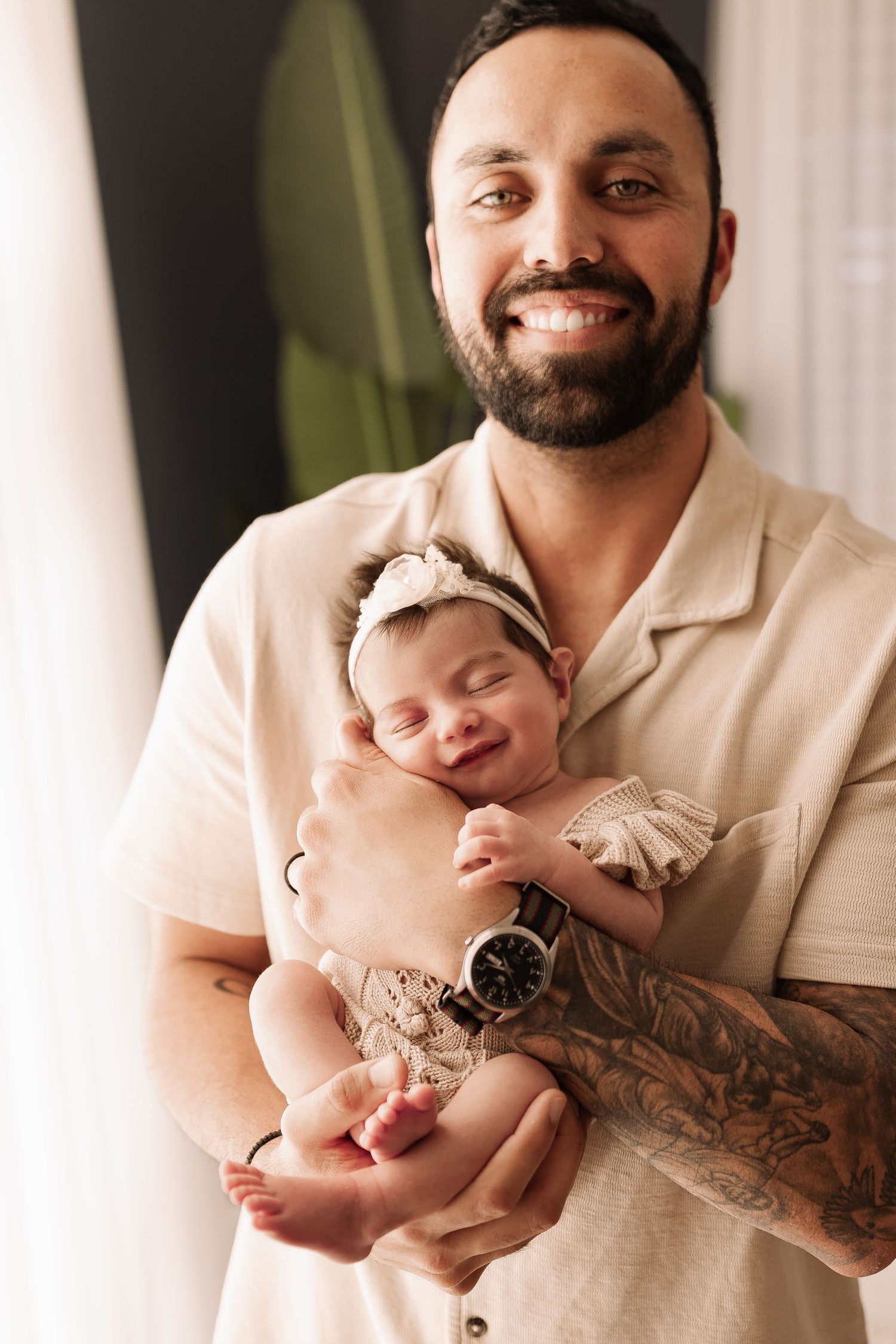 Smiling bearded father with tattoos holding sleeping newborn baby girl in cream romper.