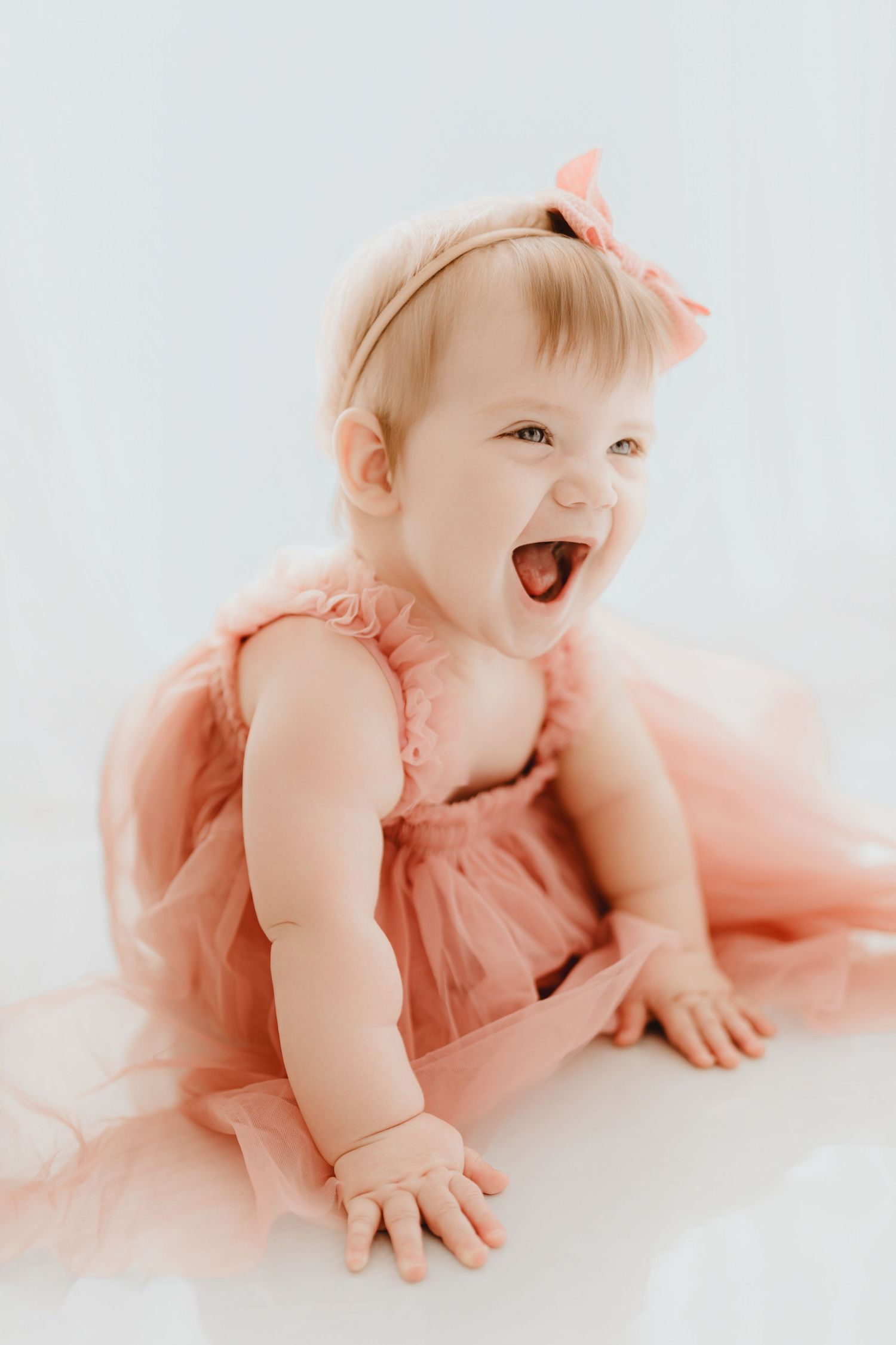 Laughing baby girl in pink tulle dress and bow headband sitting on white floor.