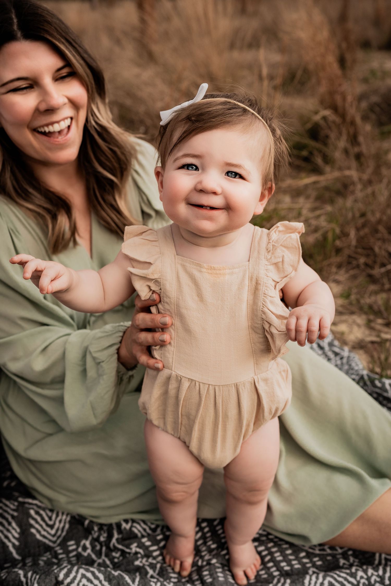 Laughing mother in green dress holding smiling baby girl in beige ruffle romper standing on a blanket outdoors.