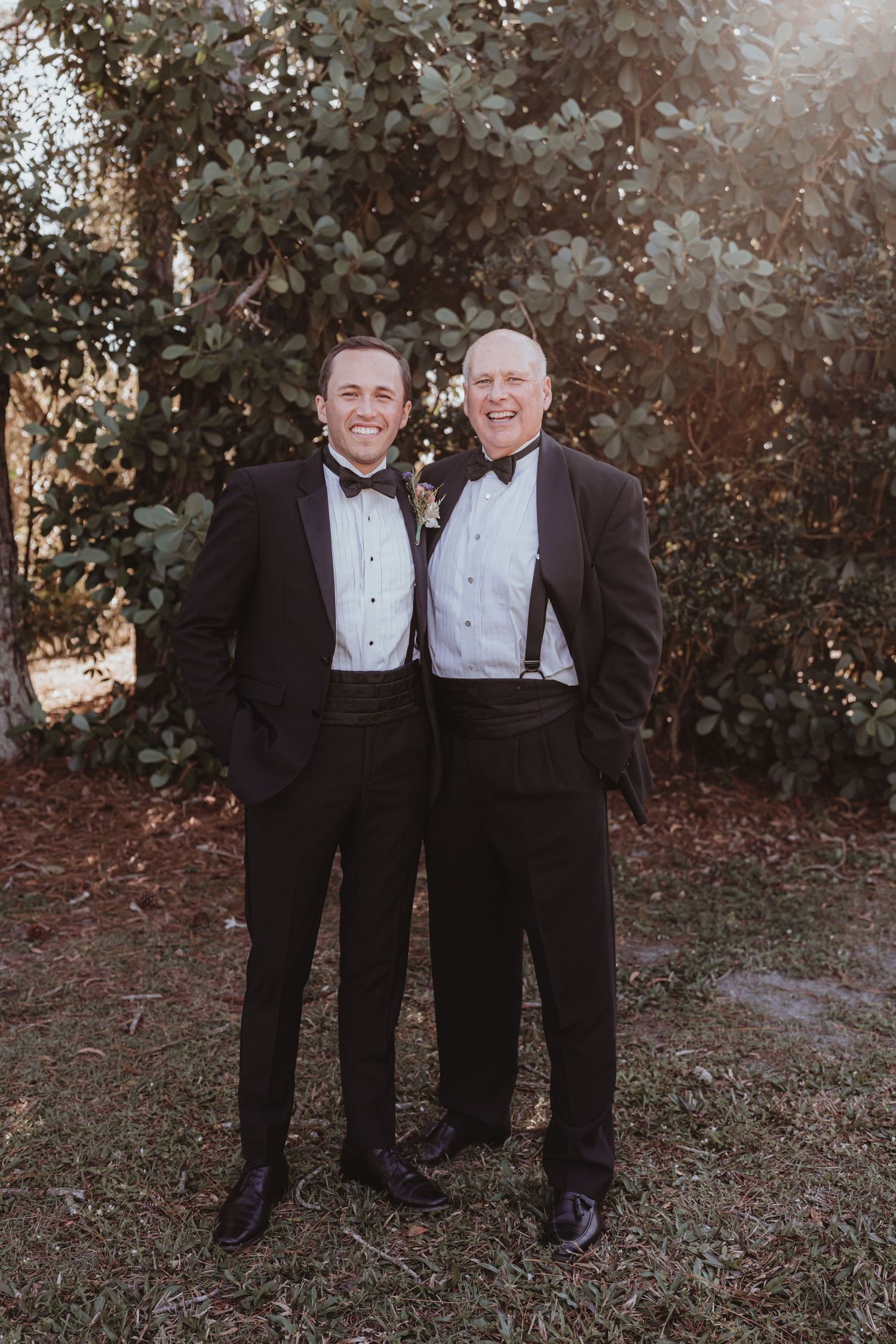 Two smiling men dressed in matching black tuxedos with bow ties pose together at a wedding.