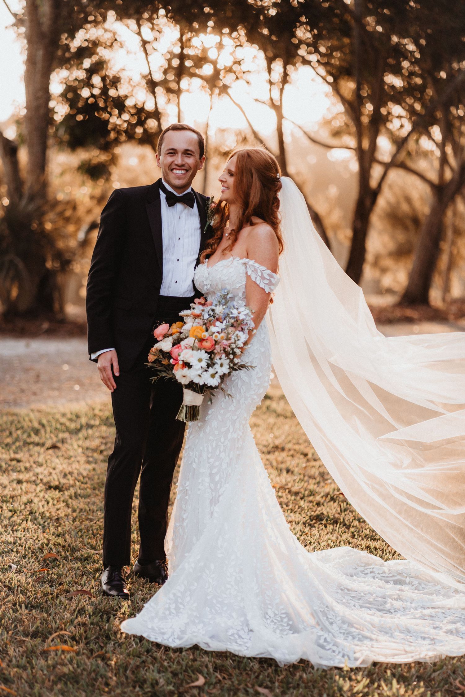 Bride and groom pose outdoors at golden hour, her lace gown and veil flowing in the breeze as she holds a colorful bouquet.