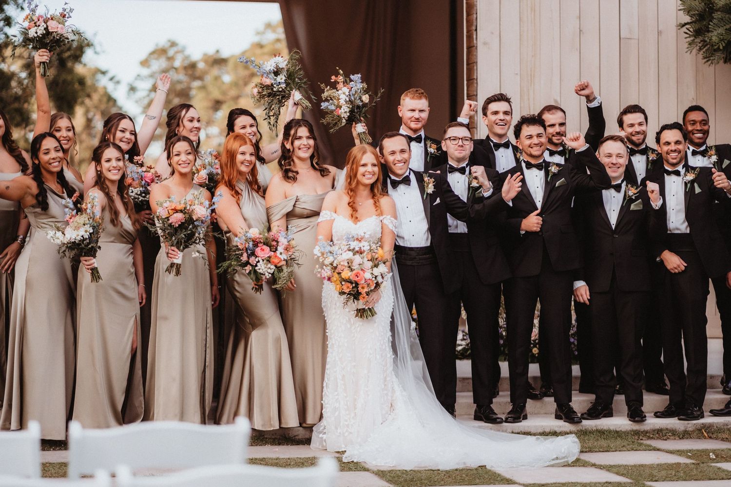 Excited wedding party celebrates outdoors, bridesmaids in champagne dresses and groomsmen in black tuxedos cheering together.