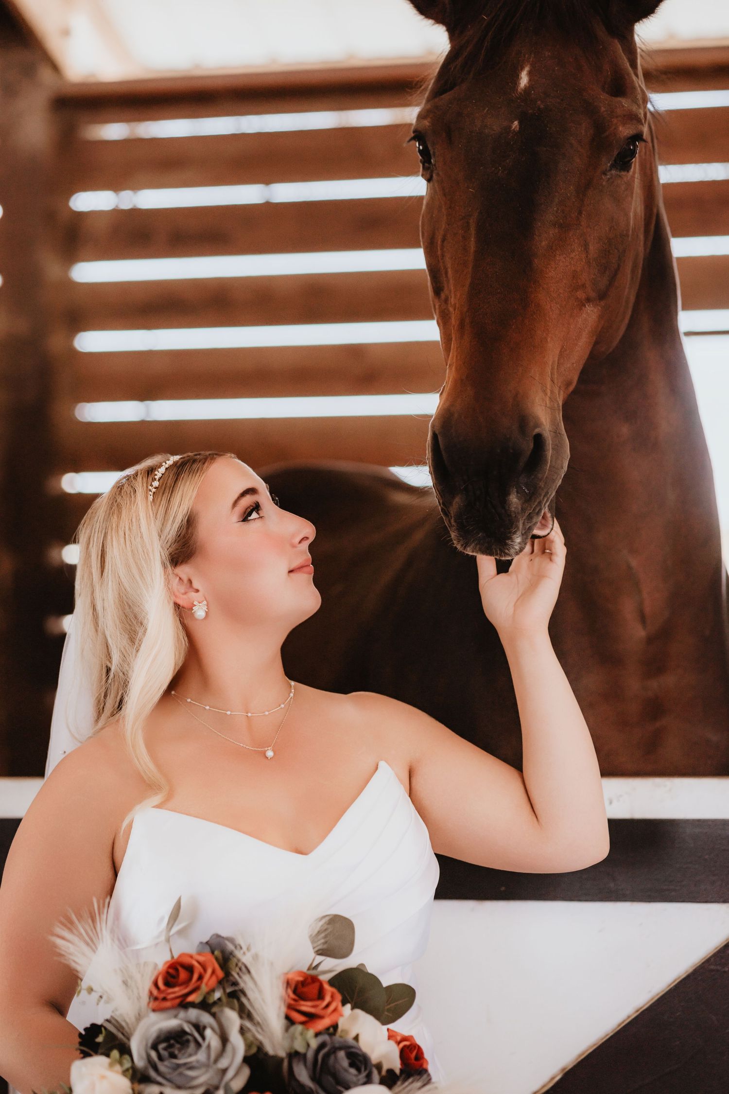 Bride in strapless white gown reaches up to touch a brown horse's nose in a barn.