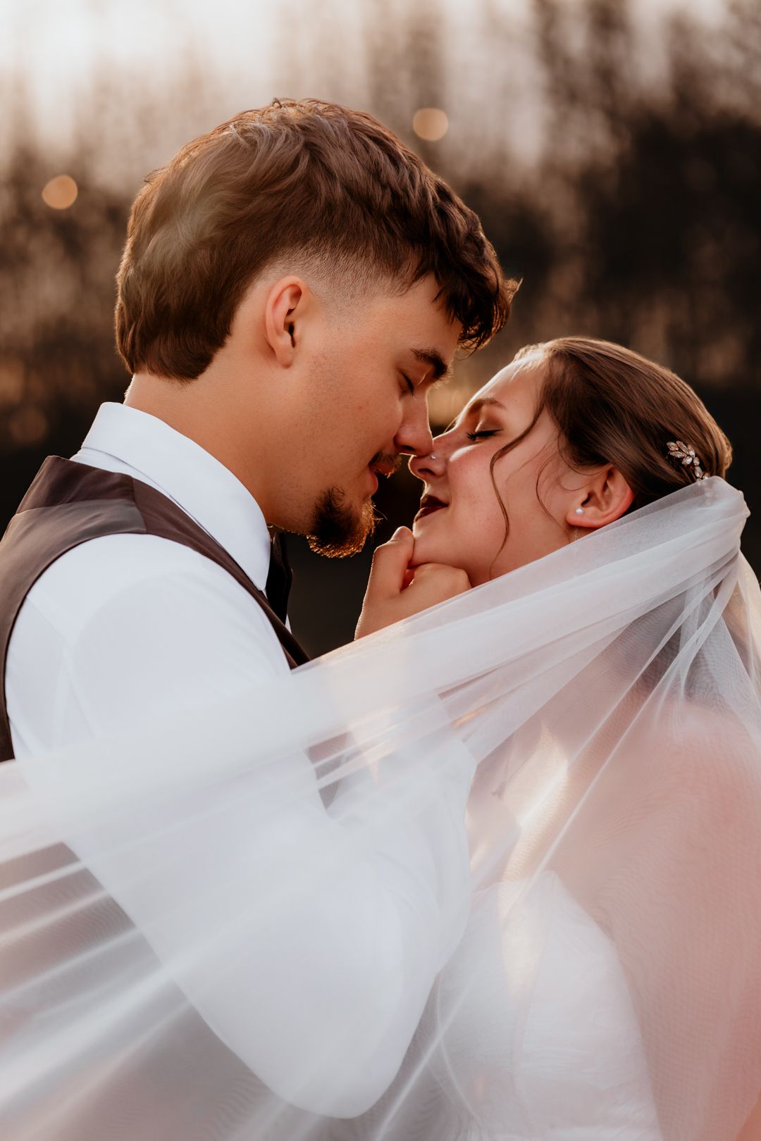 Romantic bride and groom almost kissing under a flowing veil in soft golden sunset light.
