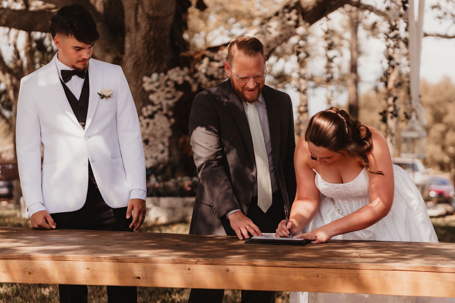 Bride signing wedding certificate at wooden table with groom in white tuxedo and officiant outdoors.