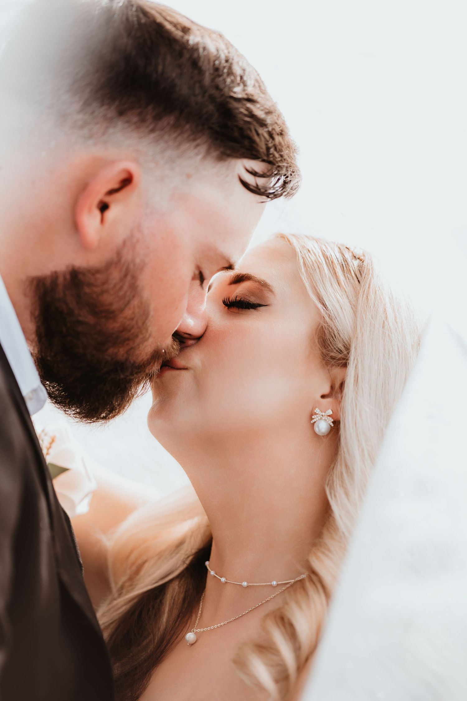 Bride and groom share an intimate kiss under a veil, bride wearing pearl earrings and a delicate necklace.