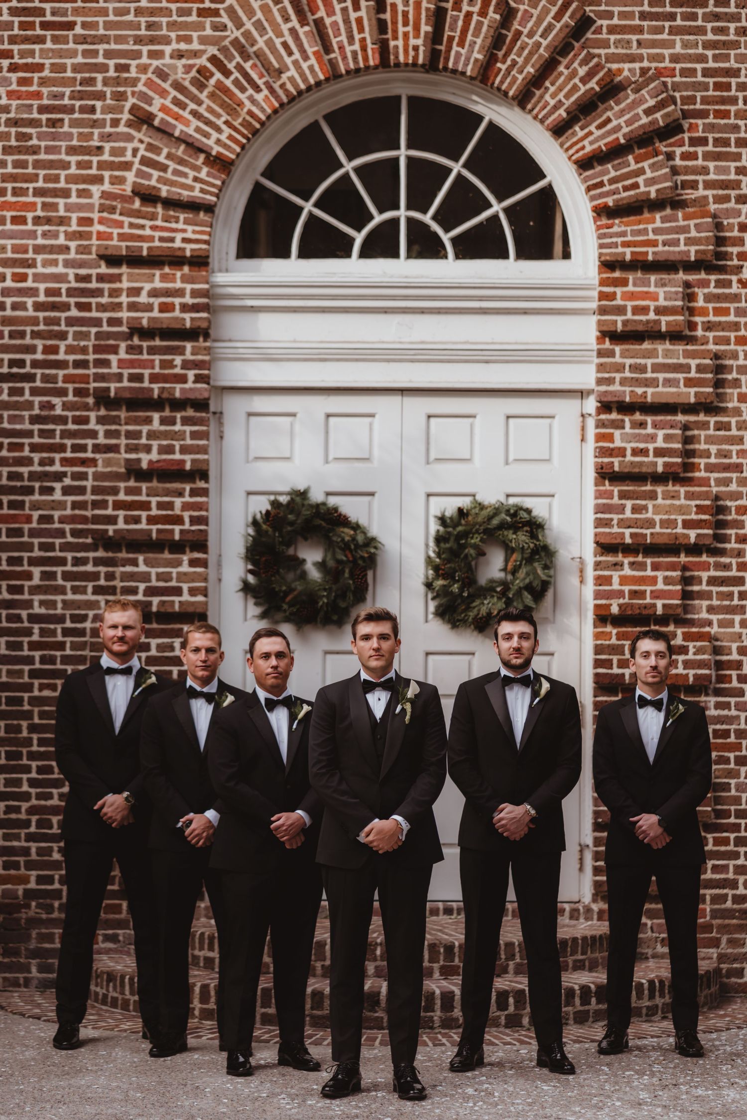 Groom and five groomsmen in black tuxedos posing formally before white arched door with holiday wreaths.