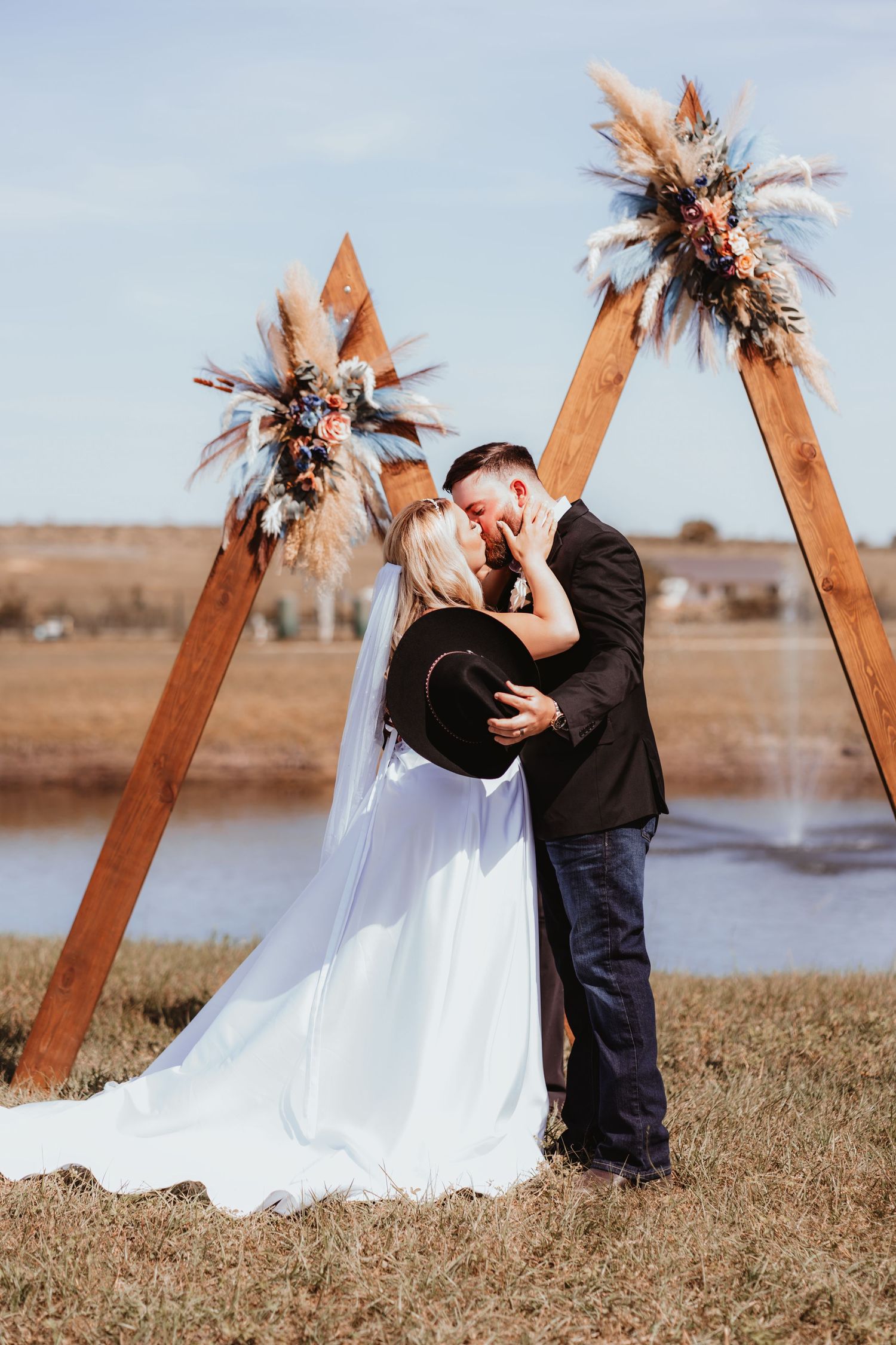 Bride and groom share first kiss under rustic wooden triangle arch adorned with pampas grass and wildflowers at outdoor wedding.