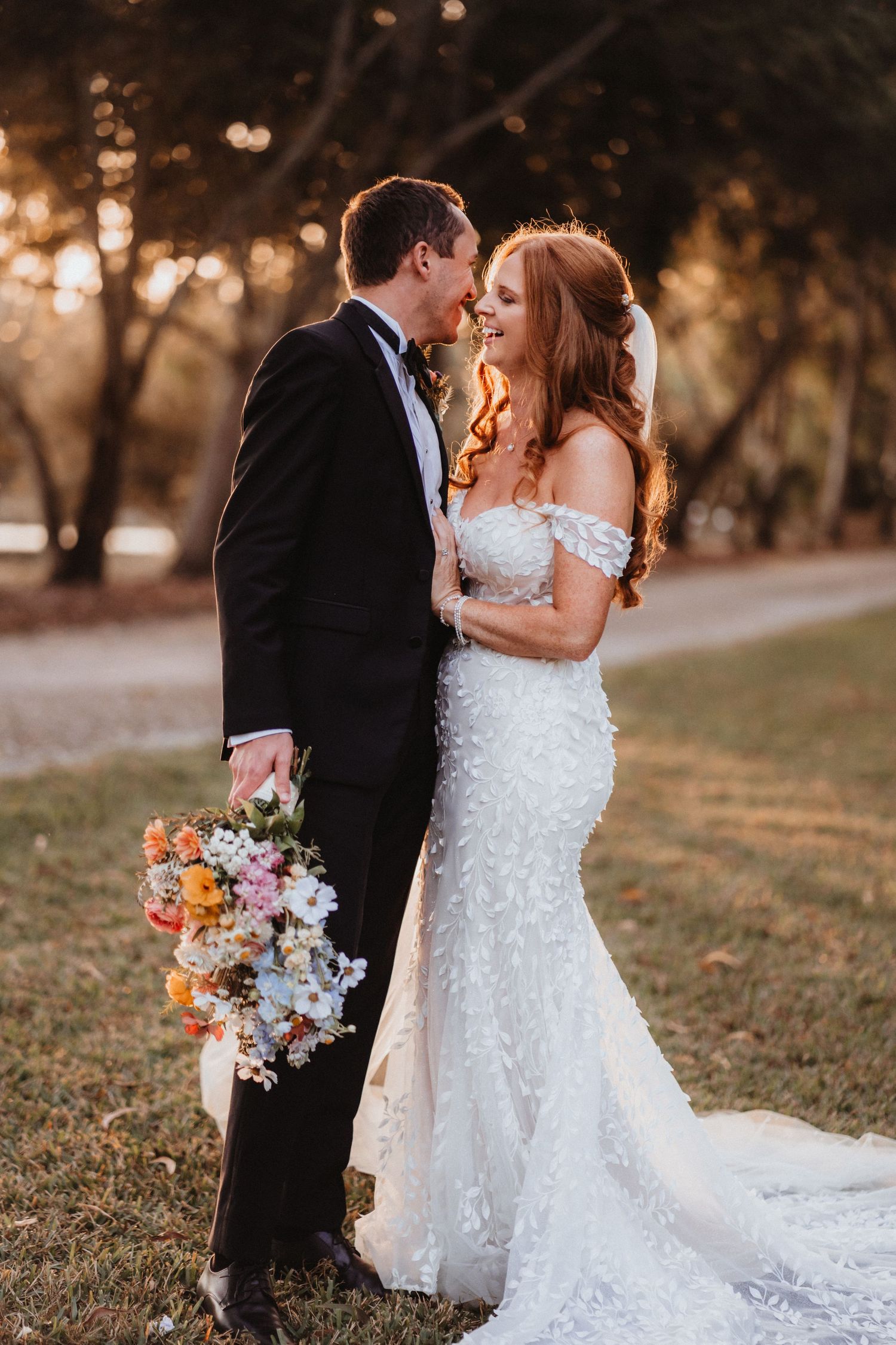 Bride and groom lean in for a kiss outdoors at sunset, bride holding vibrant mixed floral bouquet.