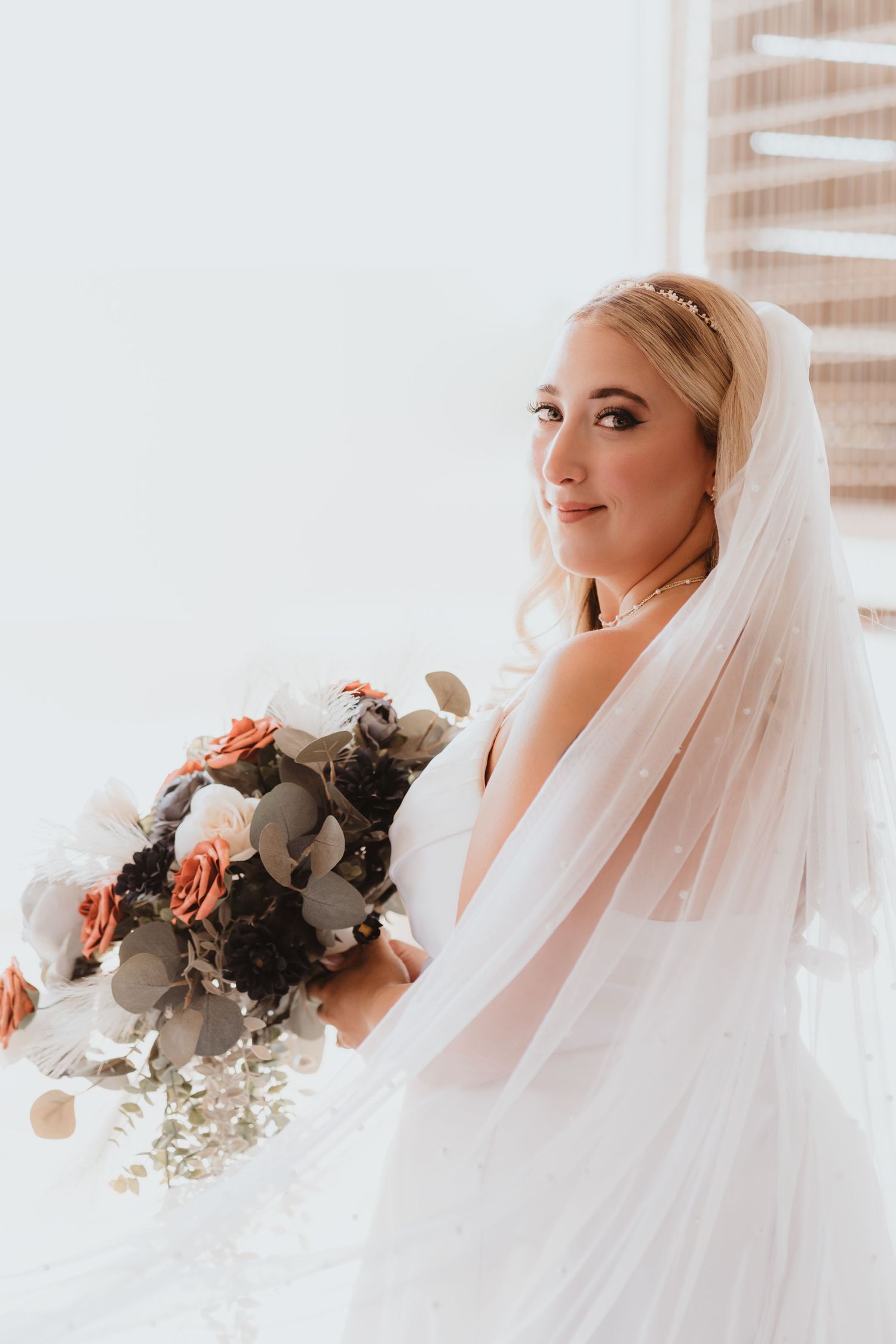Beautiful blonde bride in white gown holds bouquet of terracotta and dusty blue roses with veil.