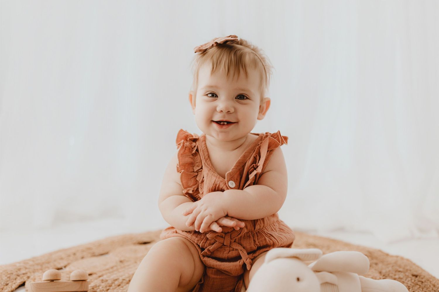 Adorable baby girl in terracotta ruffle romper with bow headband sitting on a round woven rug.