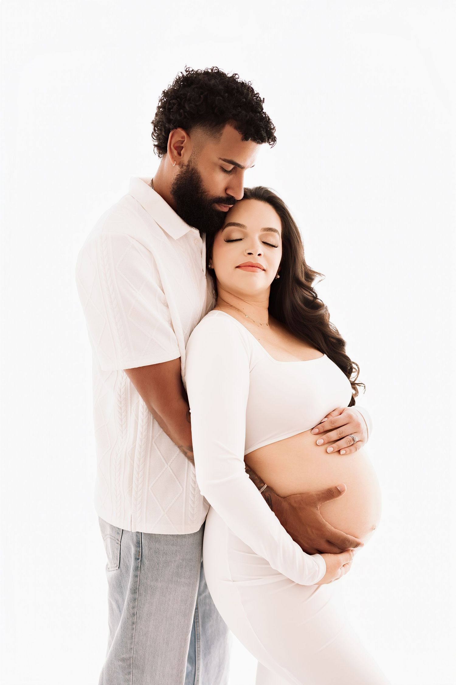 Tender maternity photo of a couple embracing, pregnant woman in white outfit cradling her baby bump against white background.