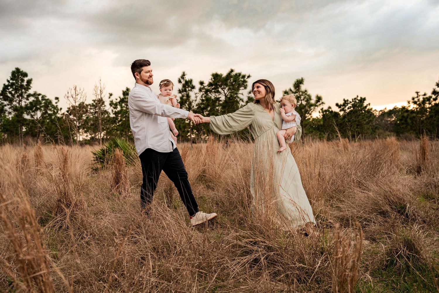 Joyful family of four walking hand-in-hand through tall golden grass at sunset in a scenic outdoor field.