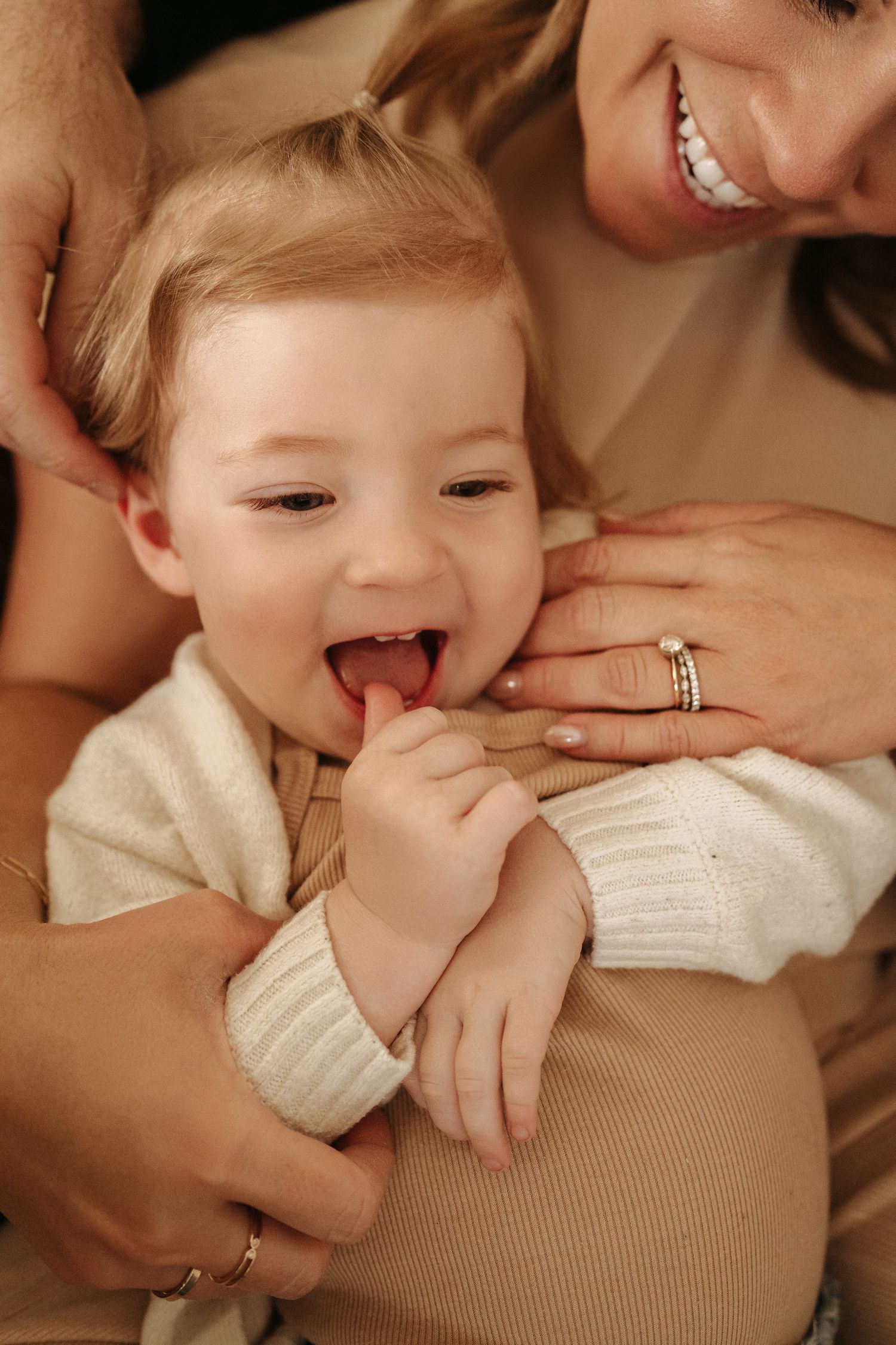 A smiling mother holds a laughing blonde toddler in a cream cardigan against her lap.