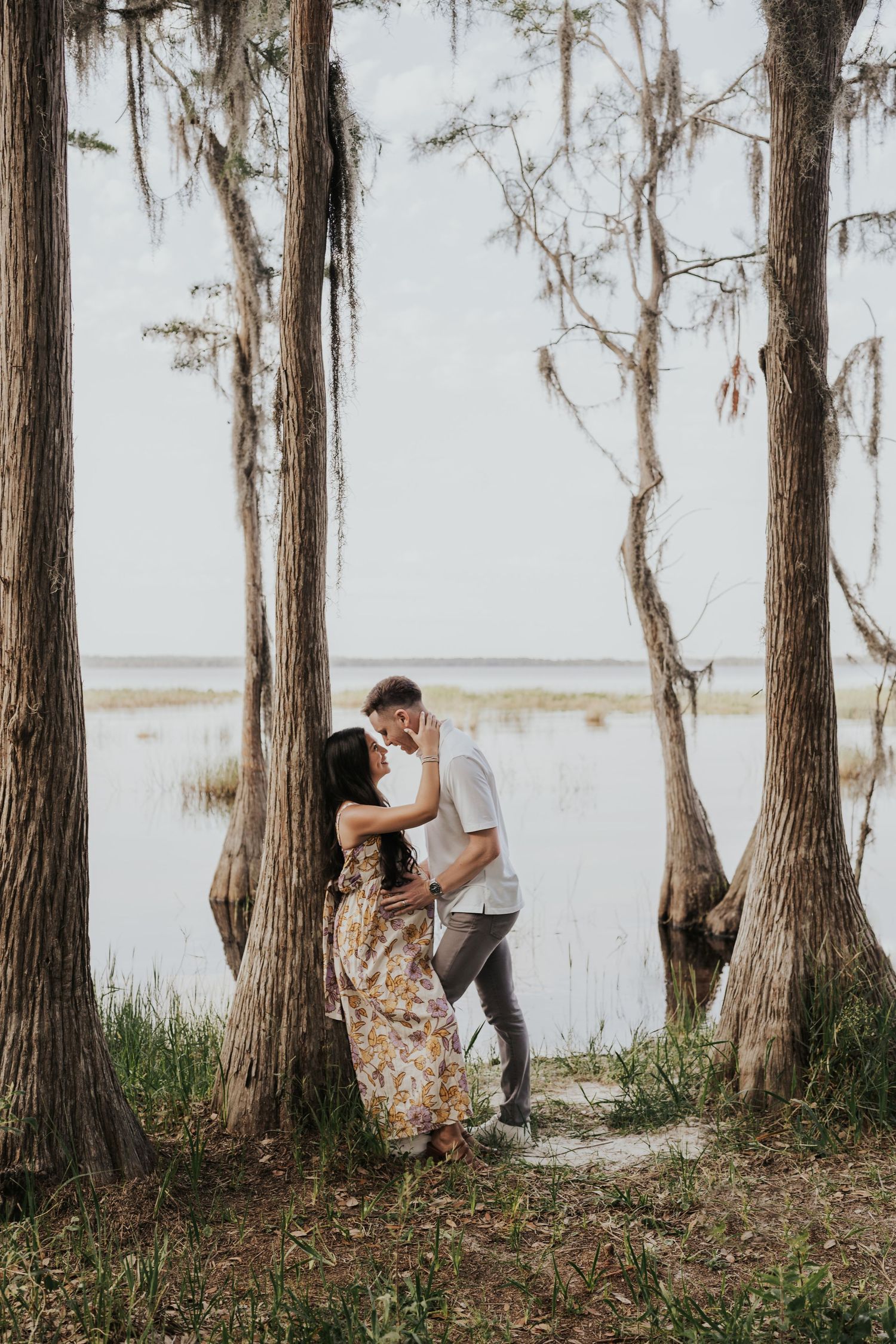 Romantic couple kissing among moss-draped cypress trees beside a serene lake in natural setting.
