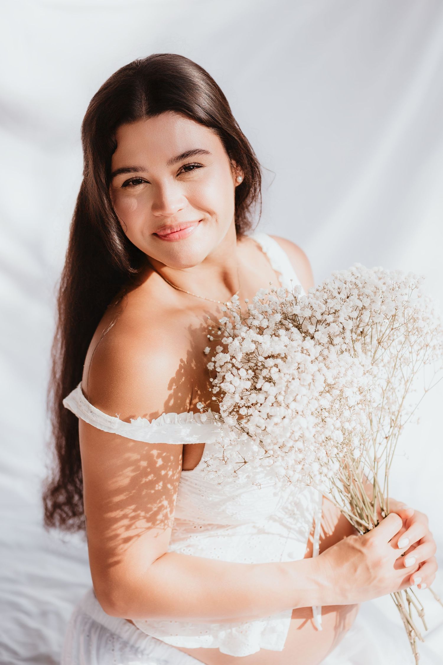 Smiling pregnant woman in white off-shoulder dress holding white wildflower bouquet indoors.