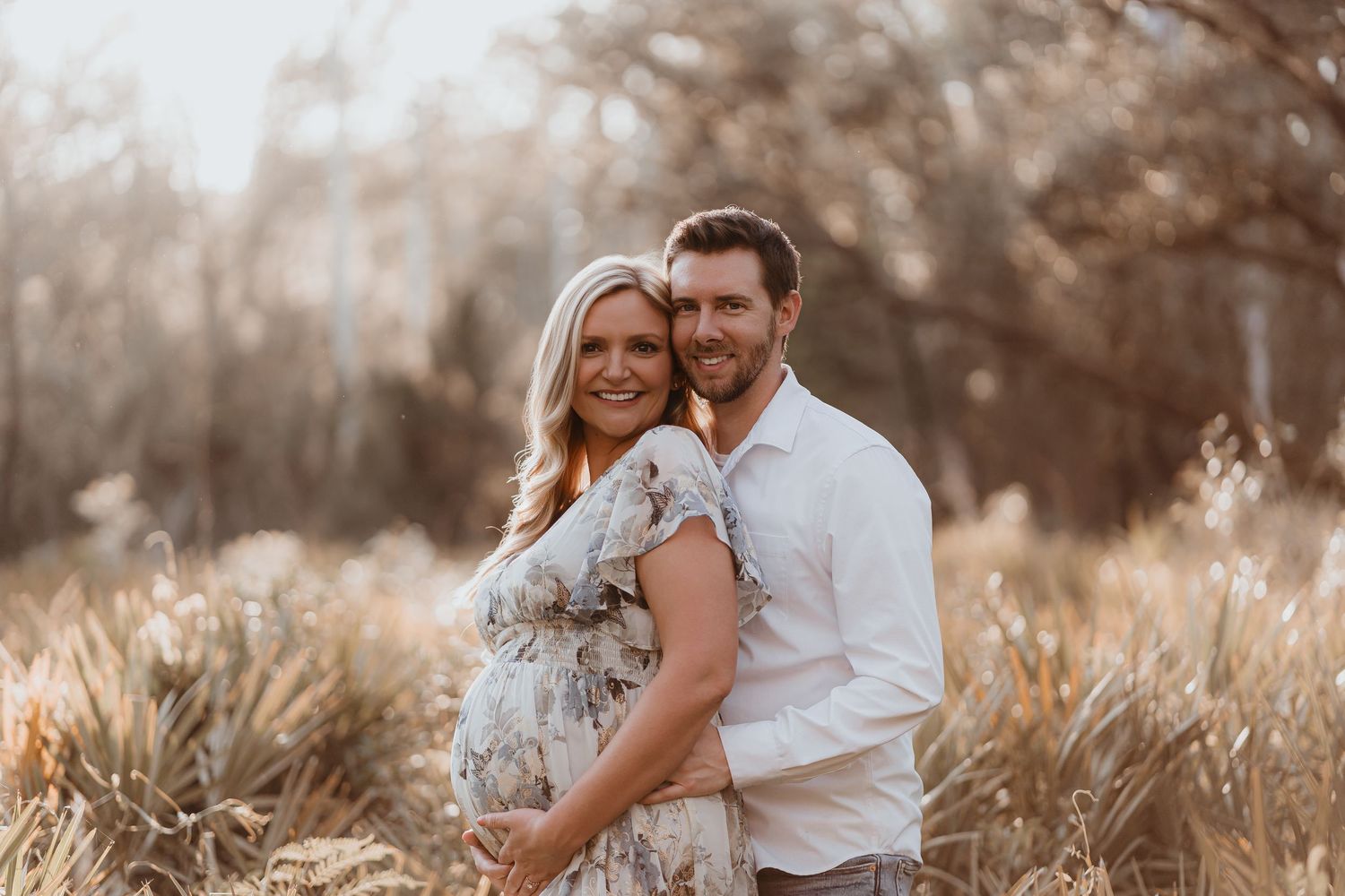 Expectant couple embraces warmly in a golden grass field during a sunny outdoor maternity photoshoot.