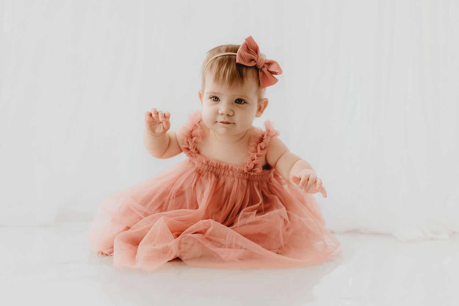Cute baby girl in pink tulle dress and bow headband sitting on white floor.