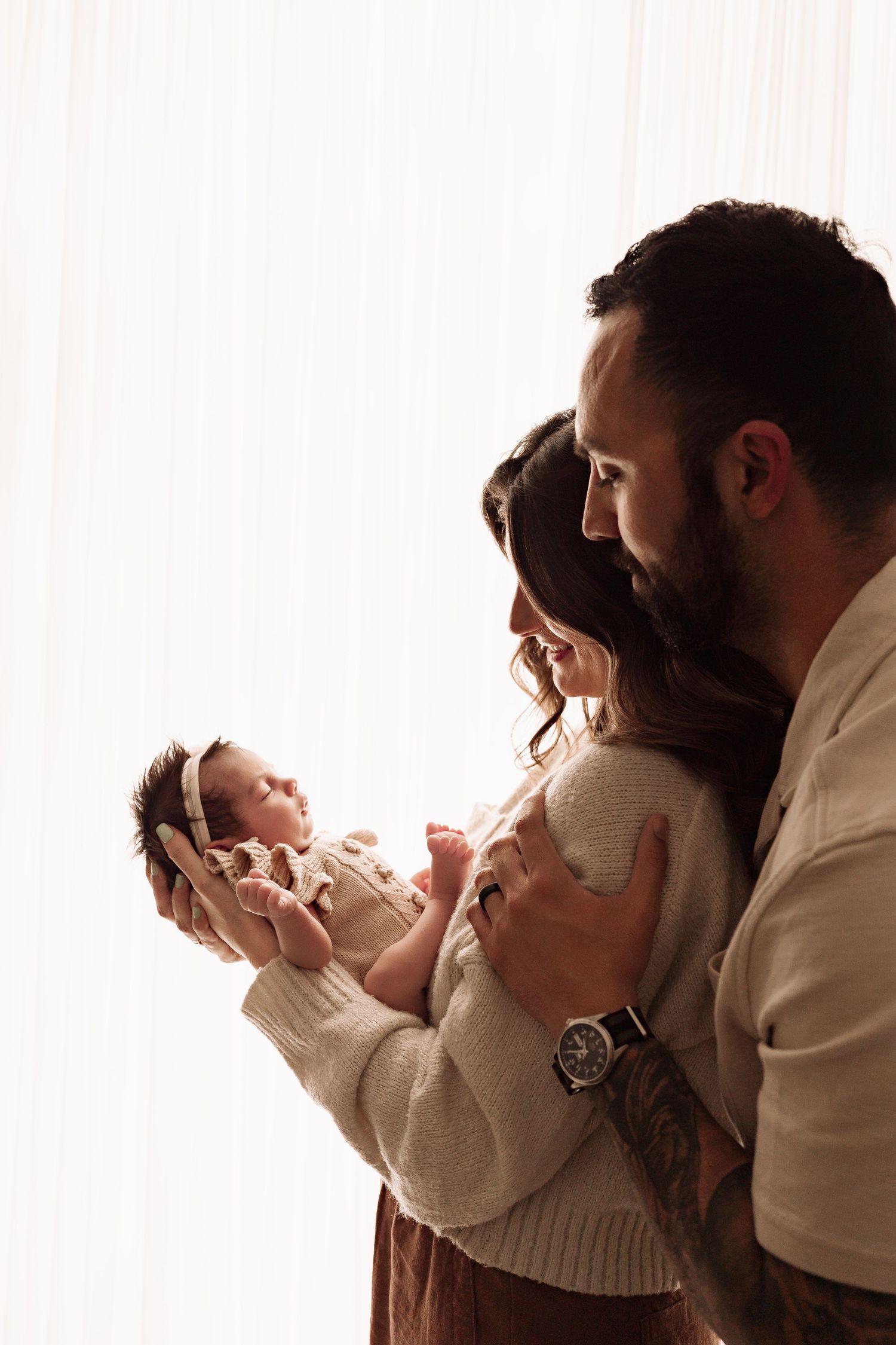 A couple stands backlit by a window, lovingly gazing at their newborn baby girl.