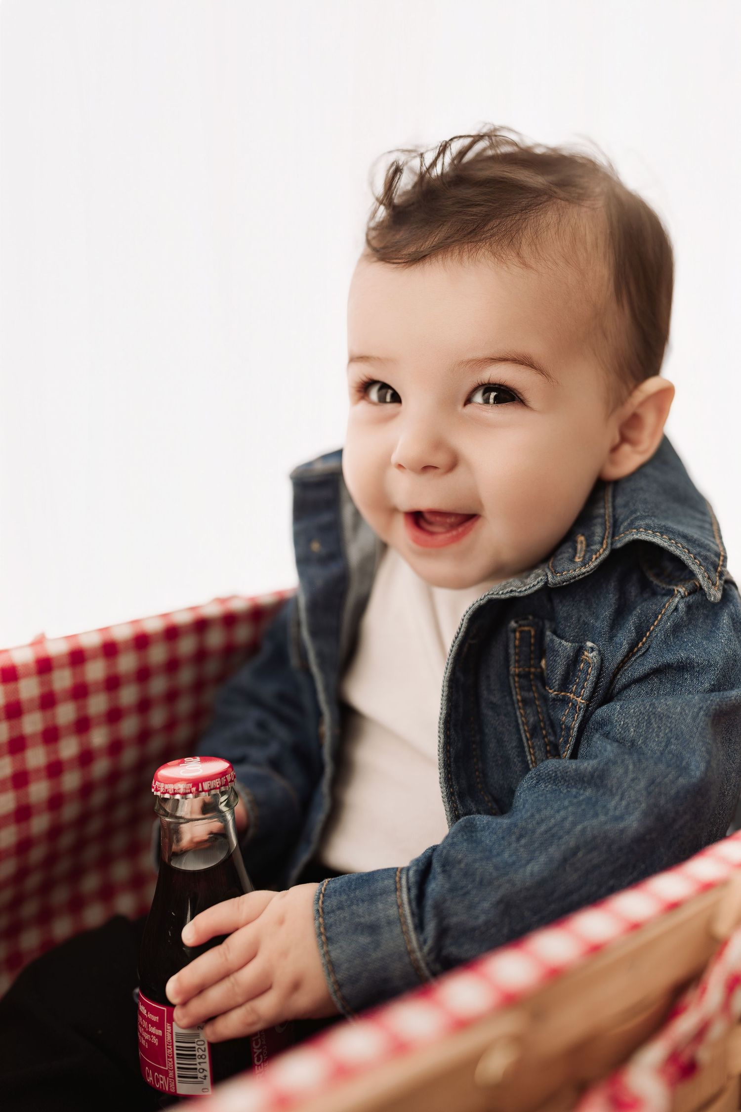 Cute baby in denim jacket smiling happily while holding a small soda bottle in a picnic basket.