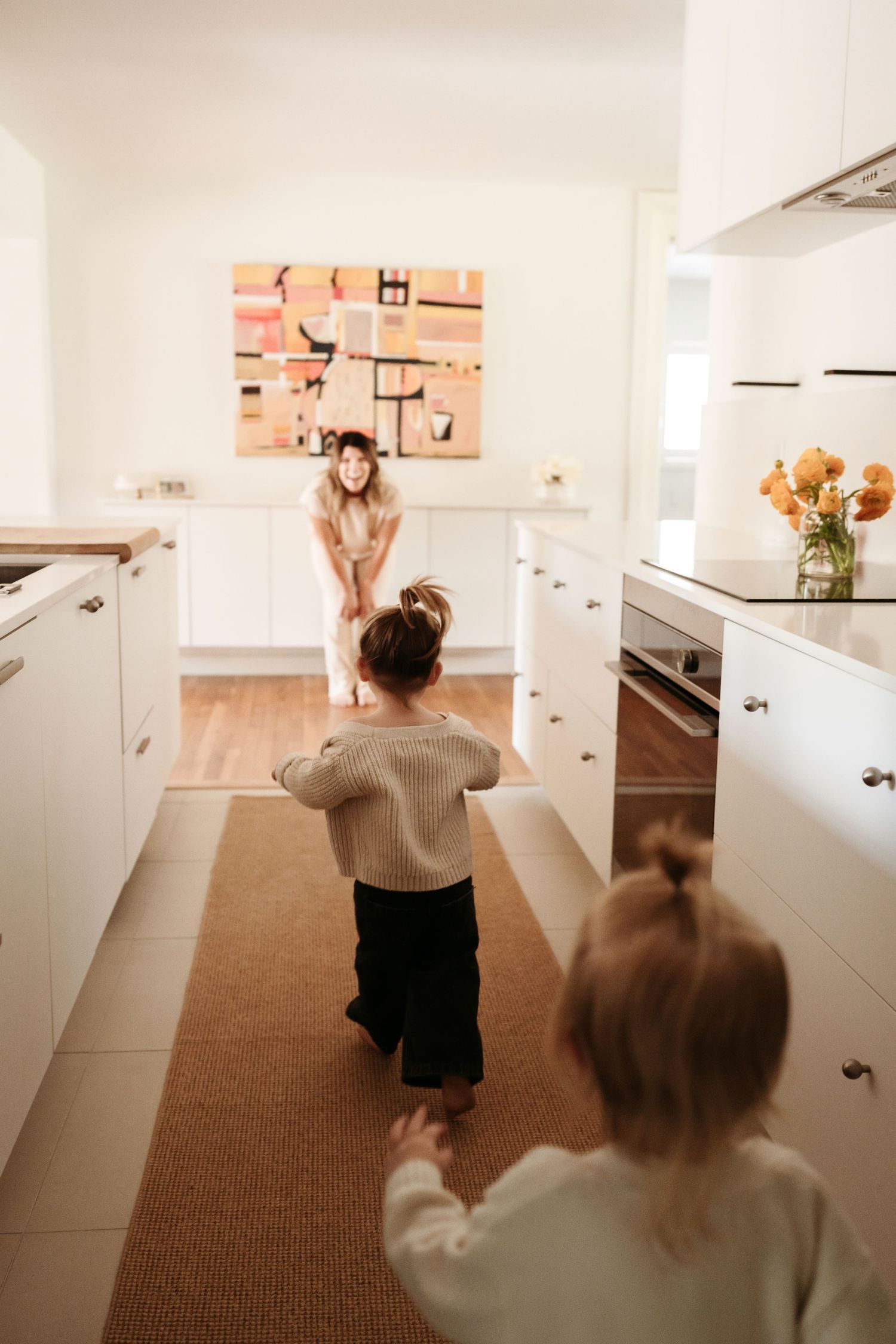 Mother crouching down in a bright white kitchen as two young children run toward her.