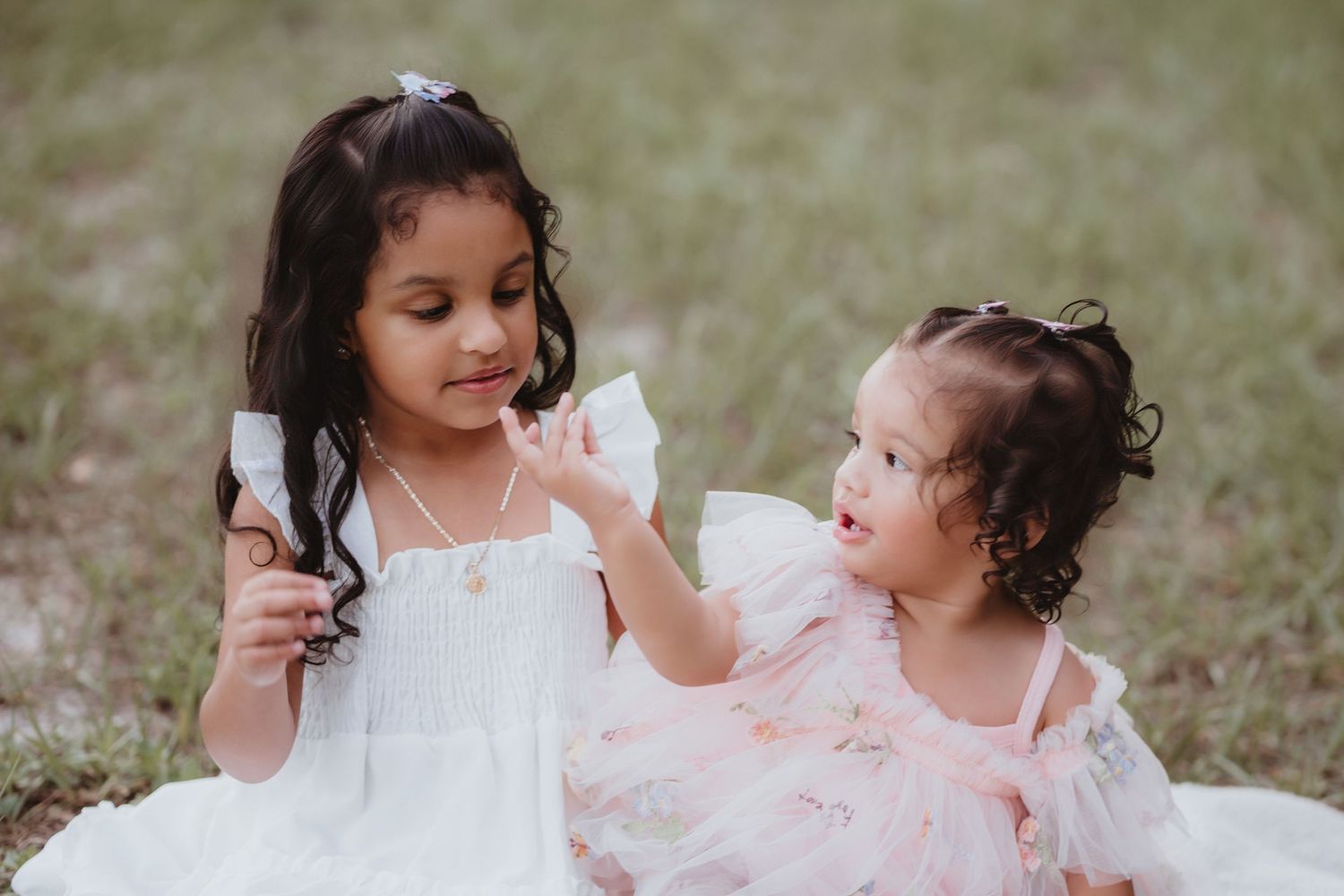 Two young sisters in white and pink dresses sitting on grass sharing a snack together.