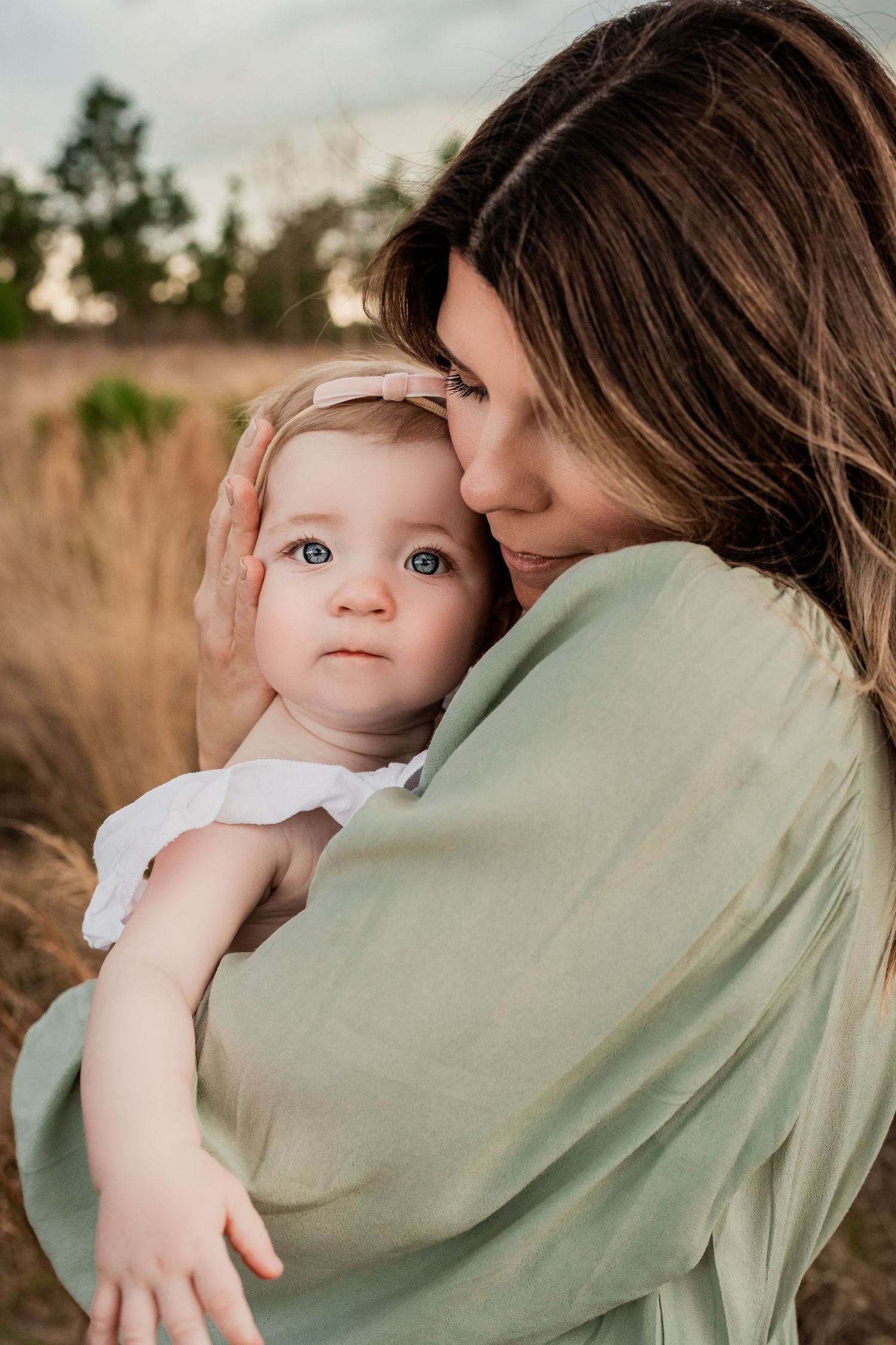 Tender moment of mother kissing baby girl's cheek while holding her in a golden grass field.