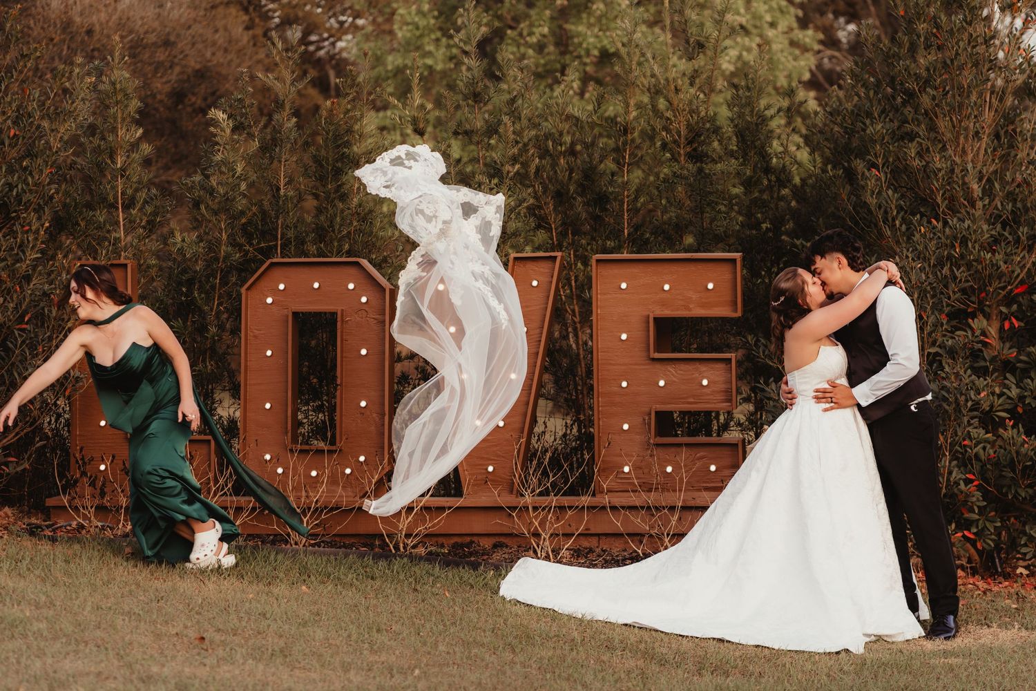 Wedding veil floating in air as bride and groom embrace near rustic LOVE marquee sign.