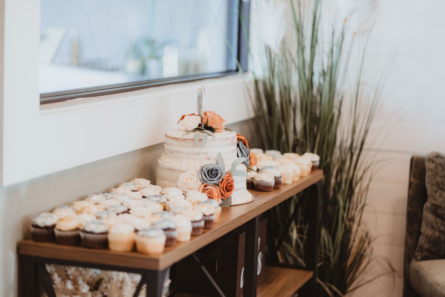 Wedding dessert table featuring tiered naked cake with floral decor and rows of frosted cupcakes near a window.