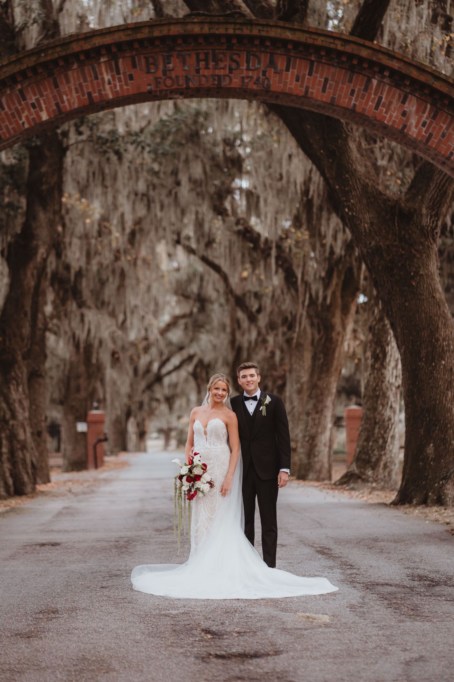 Bride and groom stand under a brick arch surrounded by moss-draped oak trees on their wedding day.