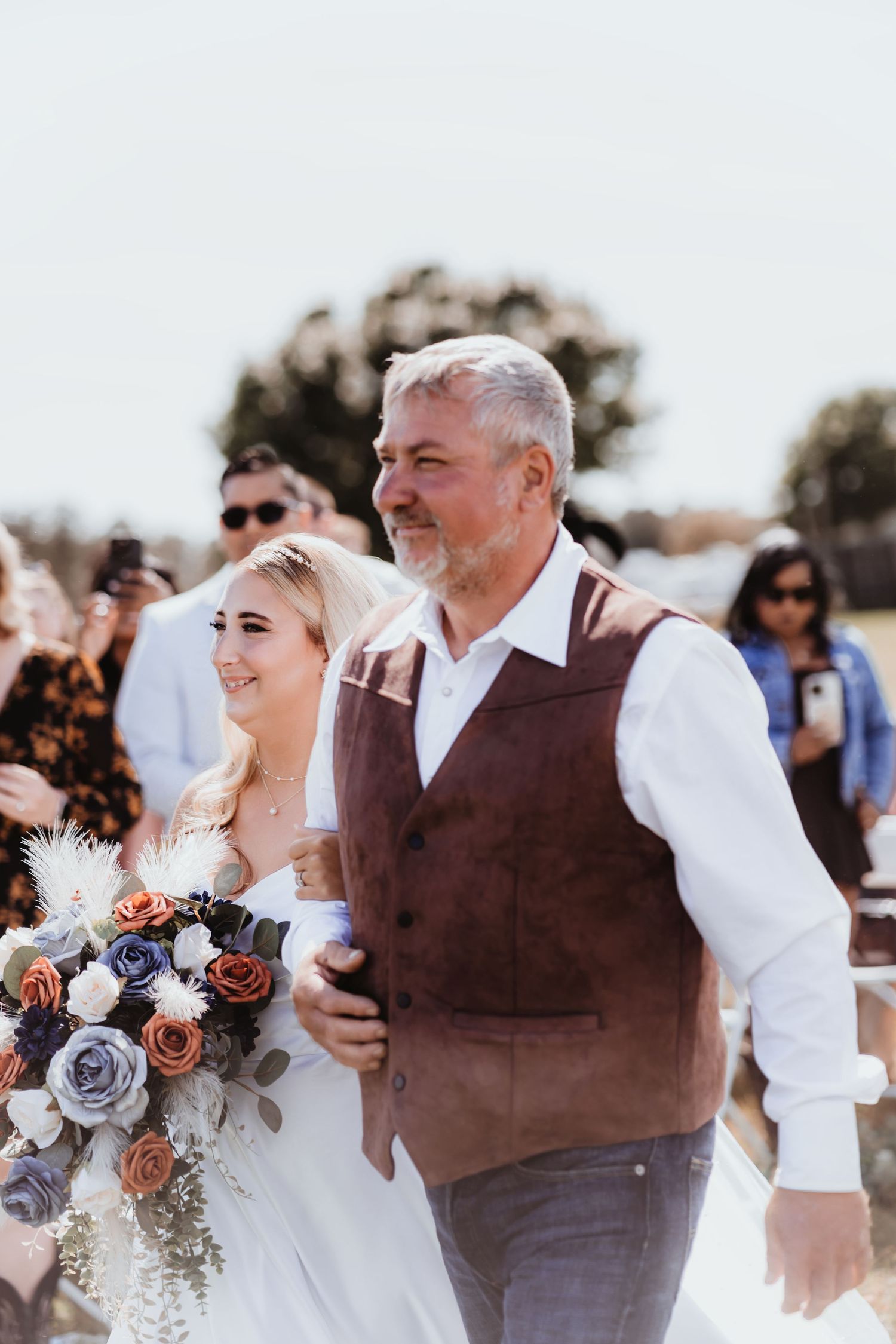 Bride with colorful boho bouquet walks down the aisle with her gray-haired father in brown vest.
