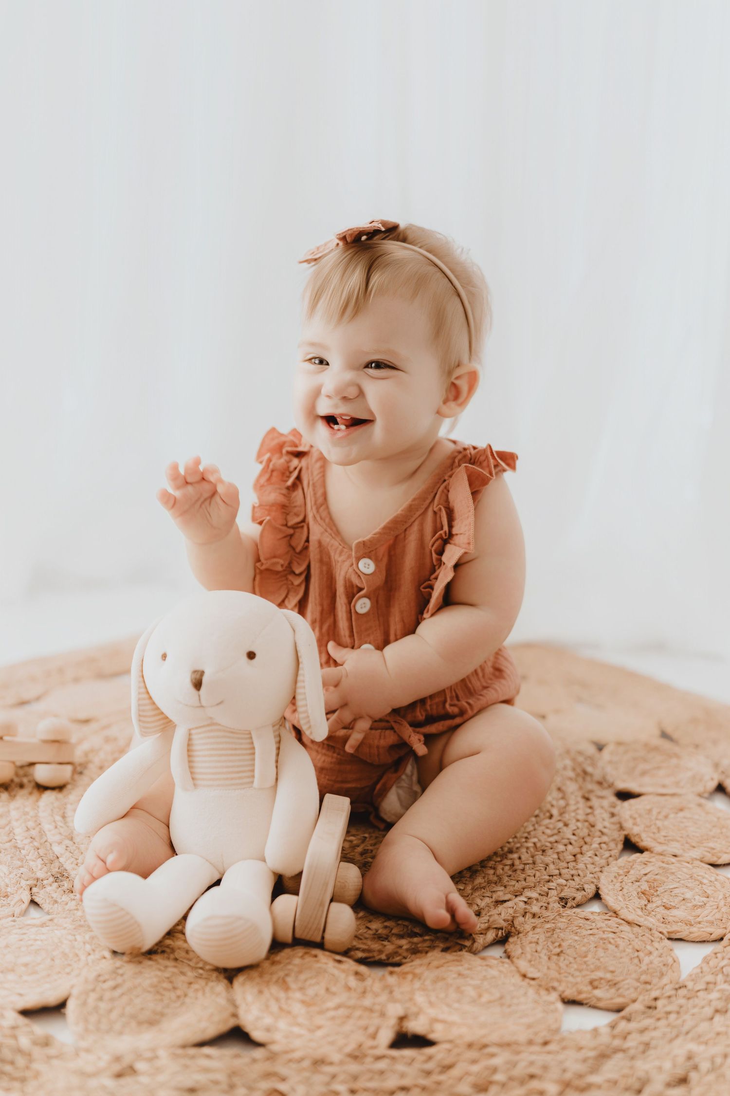 Joyful baby girl in rust ruffle romper laughing beside a white stuffed bunny on woven rug.