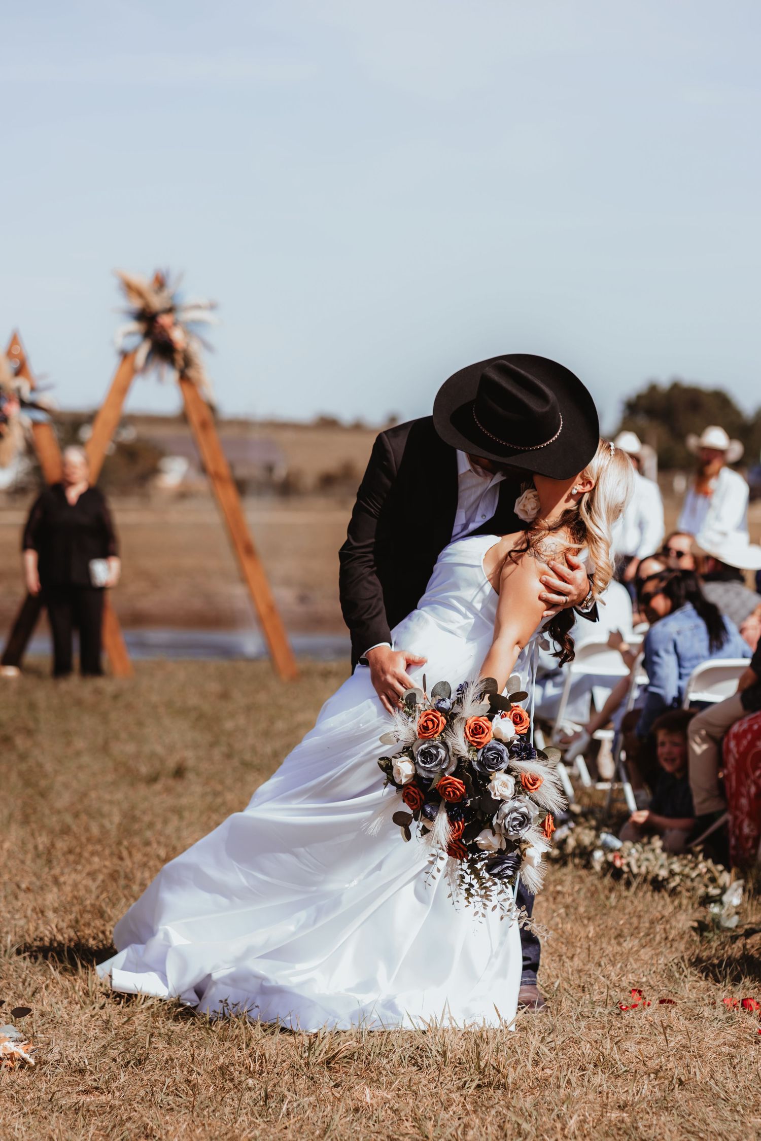 Groom in black cowboy hat dips bride for romantic kiss during outdoor rustic country wedding recessional.