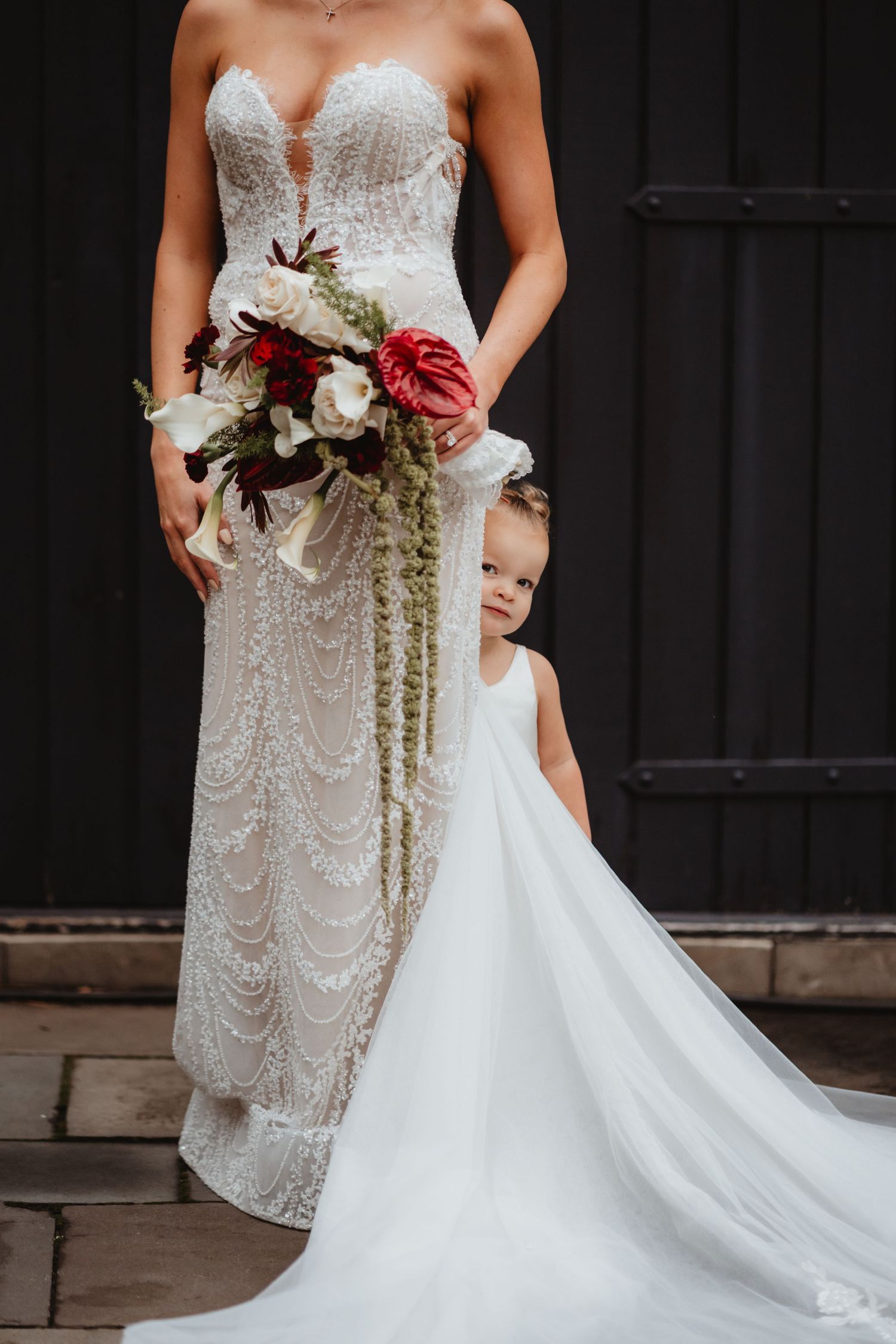 Flower girl in white dress stands beside bride holding a cascading red and white bouquet against dark wooden doors.