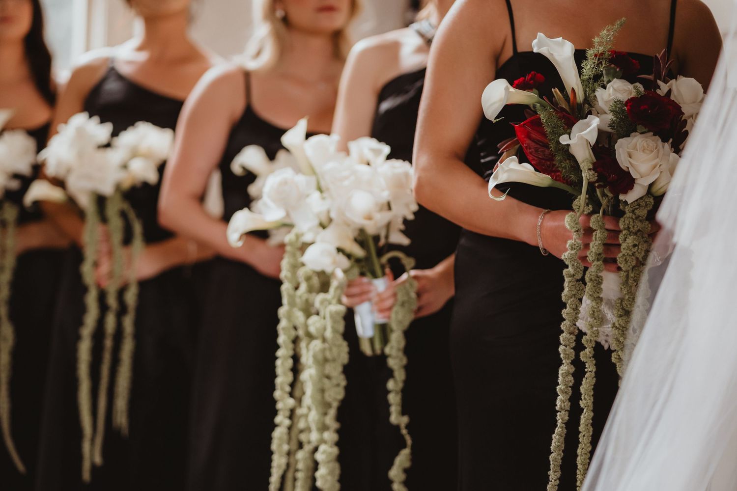 Close-up of bridesmaids in black dresses holding elegant cascading white floral bouquets with trailing greenery.
