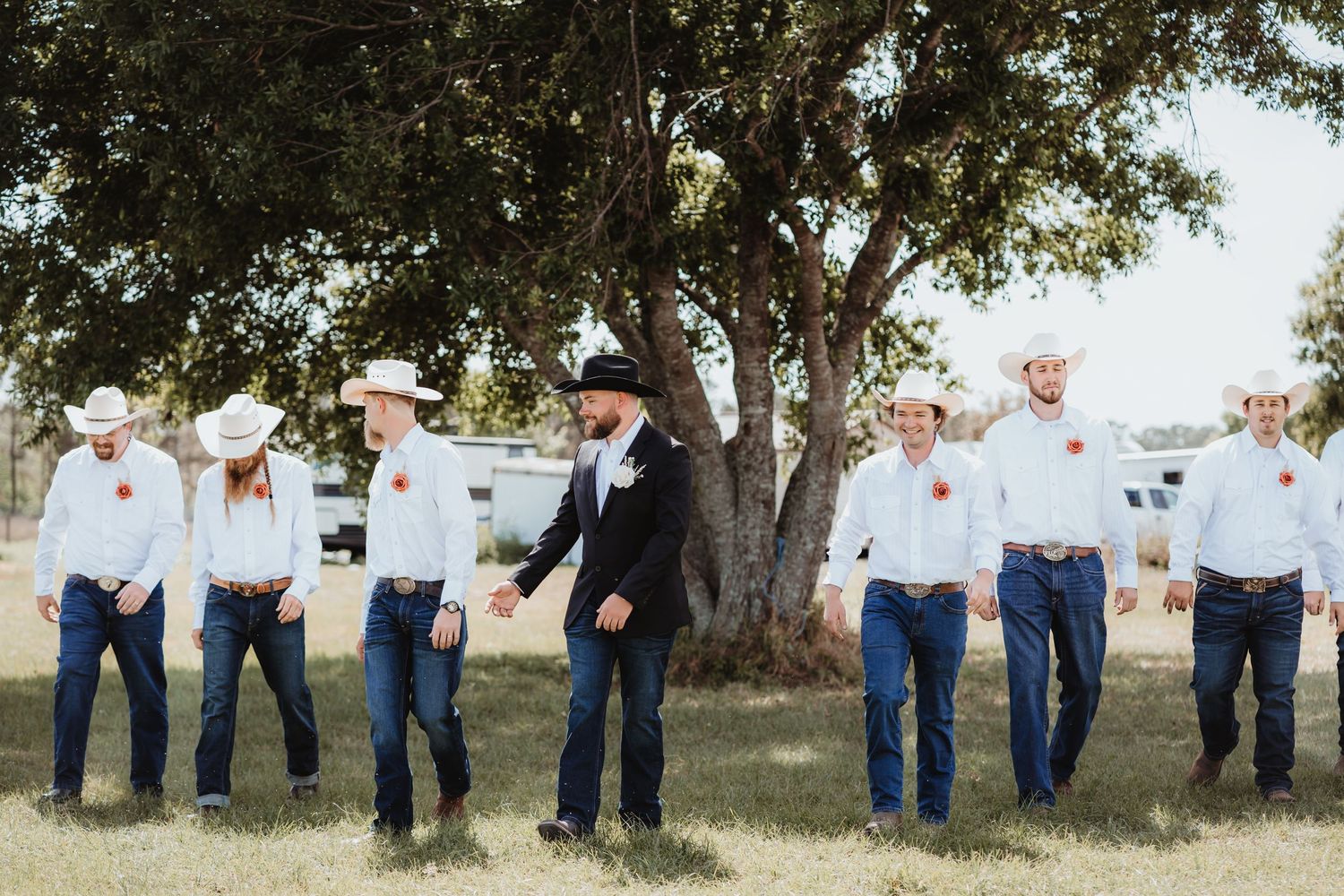Groom in black suit and cowboy hat walks with groomsmen in white shirts and cowboy hats at outdoor ranch wedding.