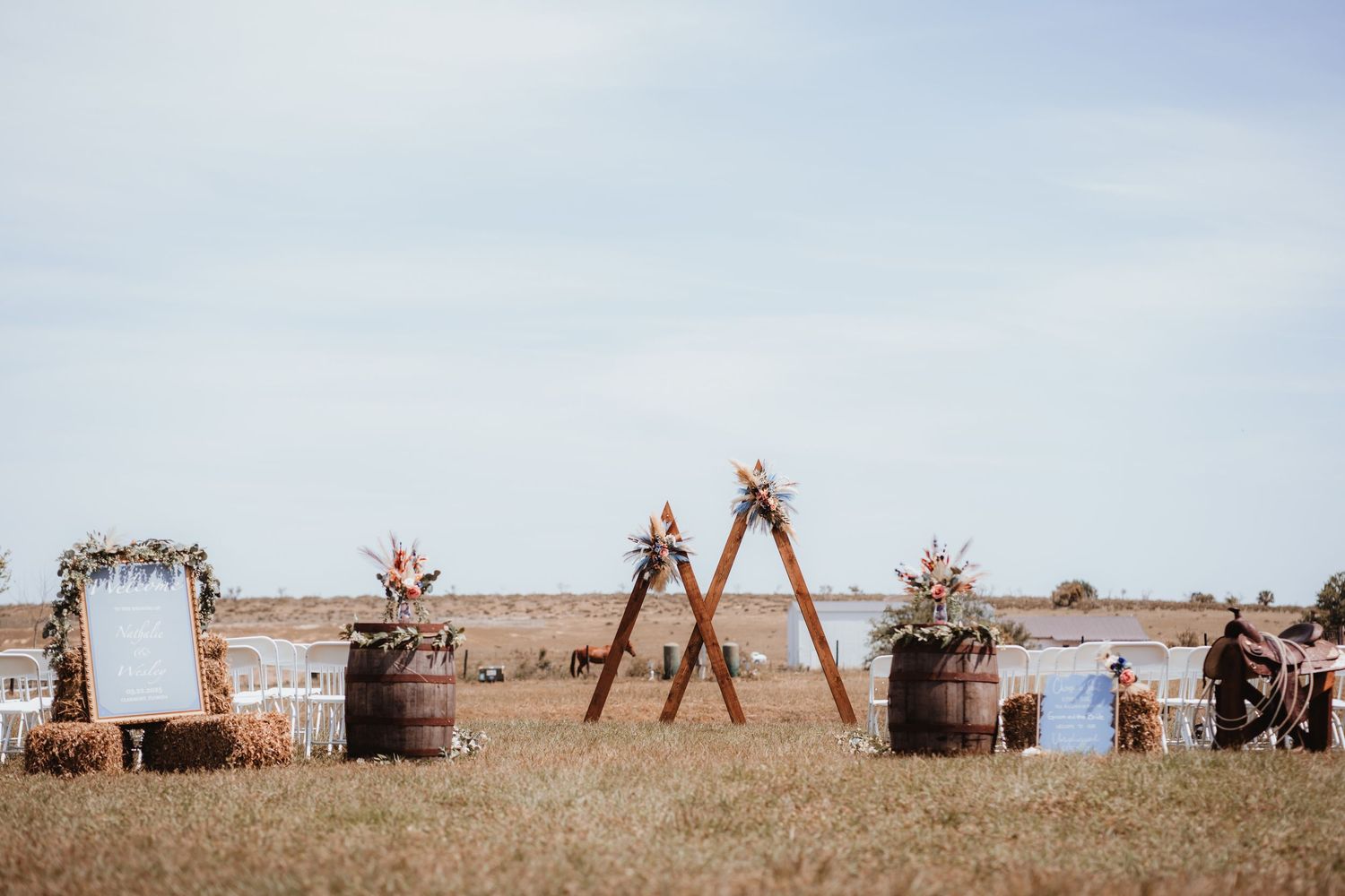 Rustic outdoor wedding ceremony setup with wooden triangle arches, barrel decorations, hay bales, and floral arrangements in an open field.