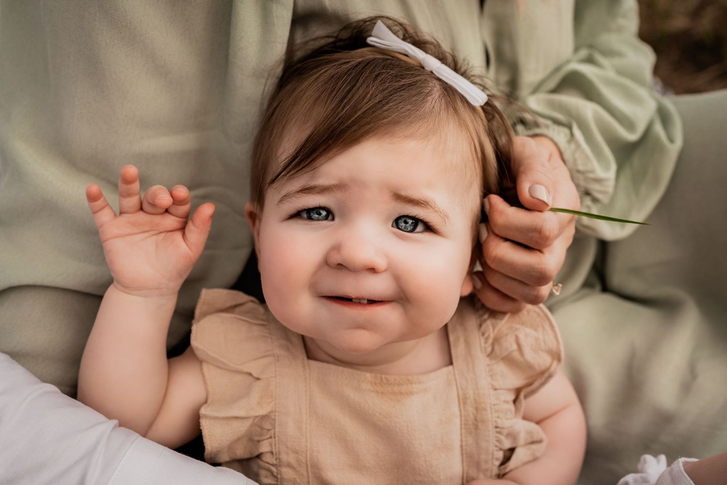 Adorable baby girl in a beige ruffle romper with a bow headband making a cute expression outdoors.