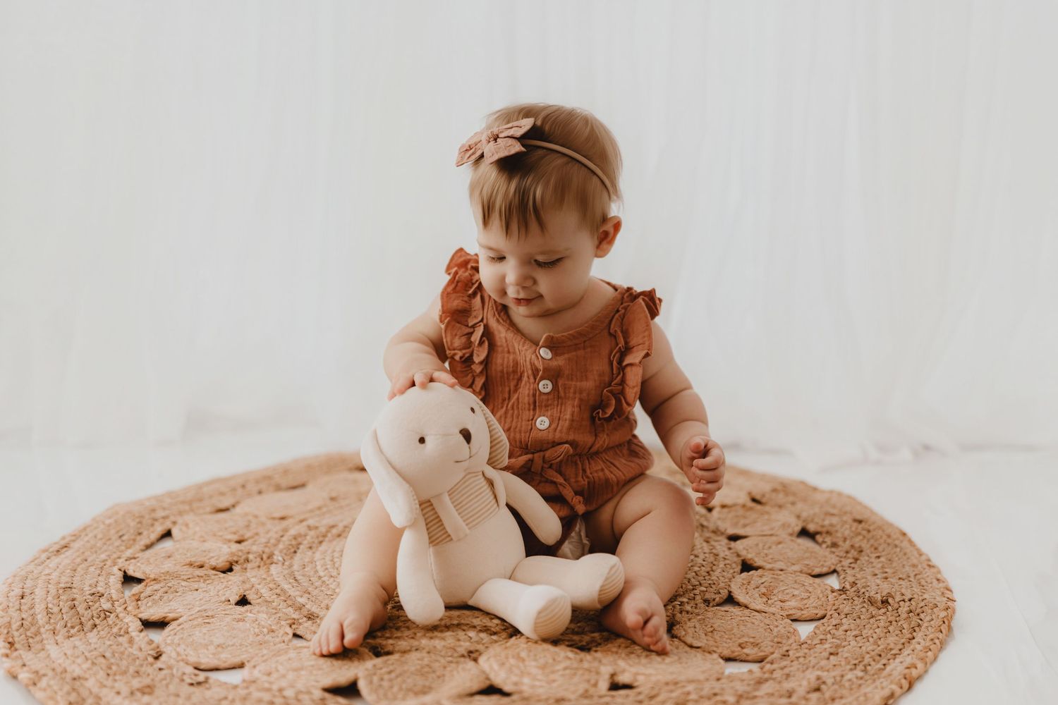 Baby girl in rust romper cuddling white stuffed bunny toy on woven rug.