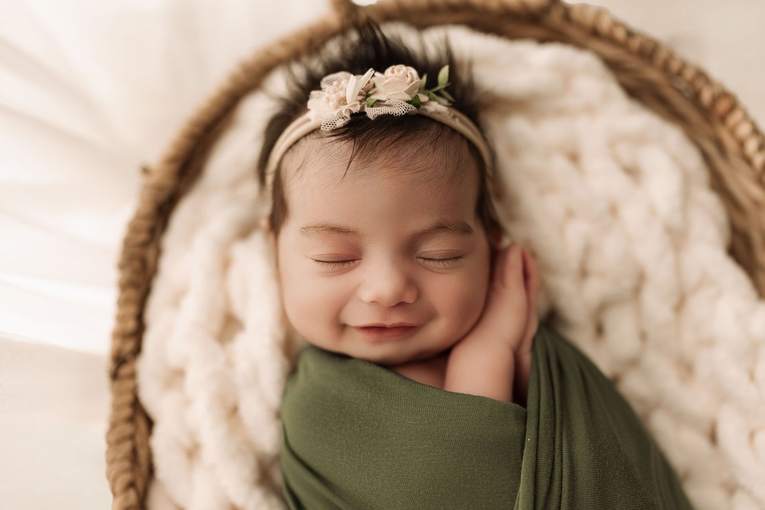 Smiling newborn baby girl in green swaddle and floral headband sleeping in cozy wicker basket.