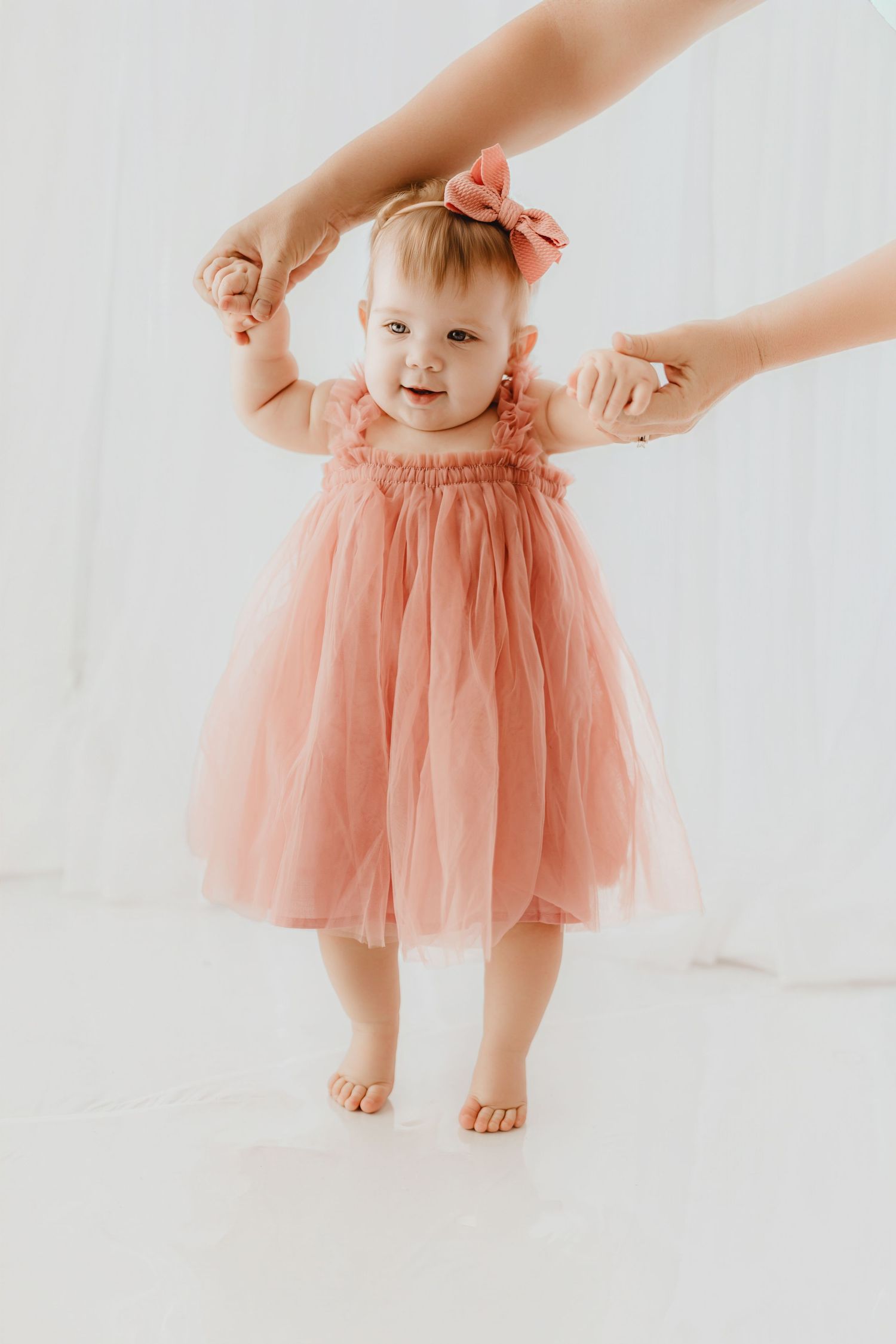 Smiling baby girl in pink tulle dress stands with adult hand support.