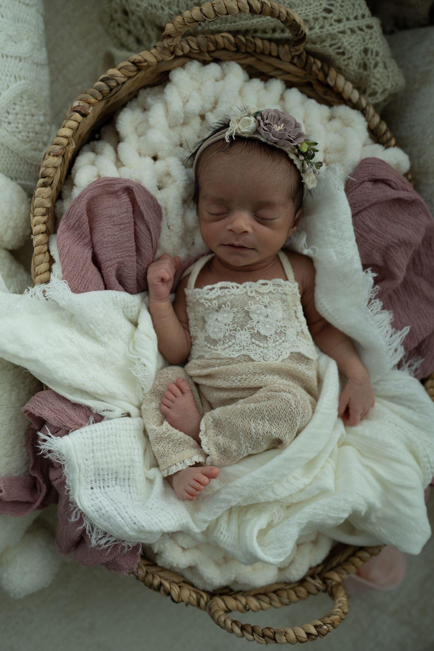 Sleeping newborn baby in lace outfit and floral headband nestled in wicker basket with white and pink wraps.