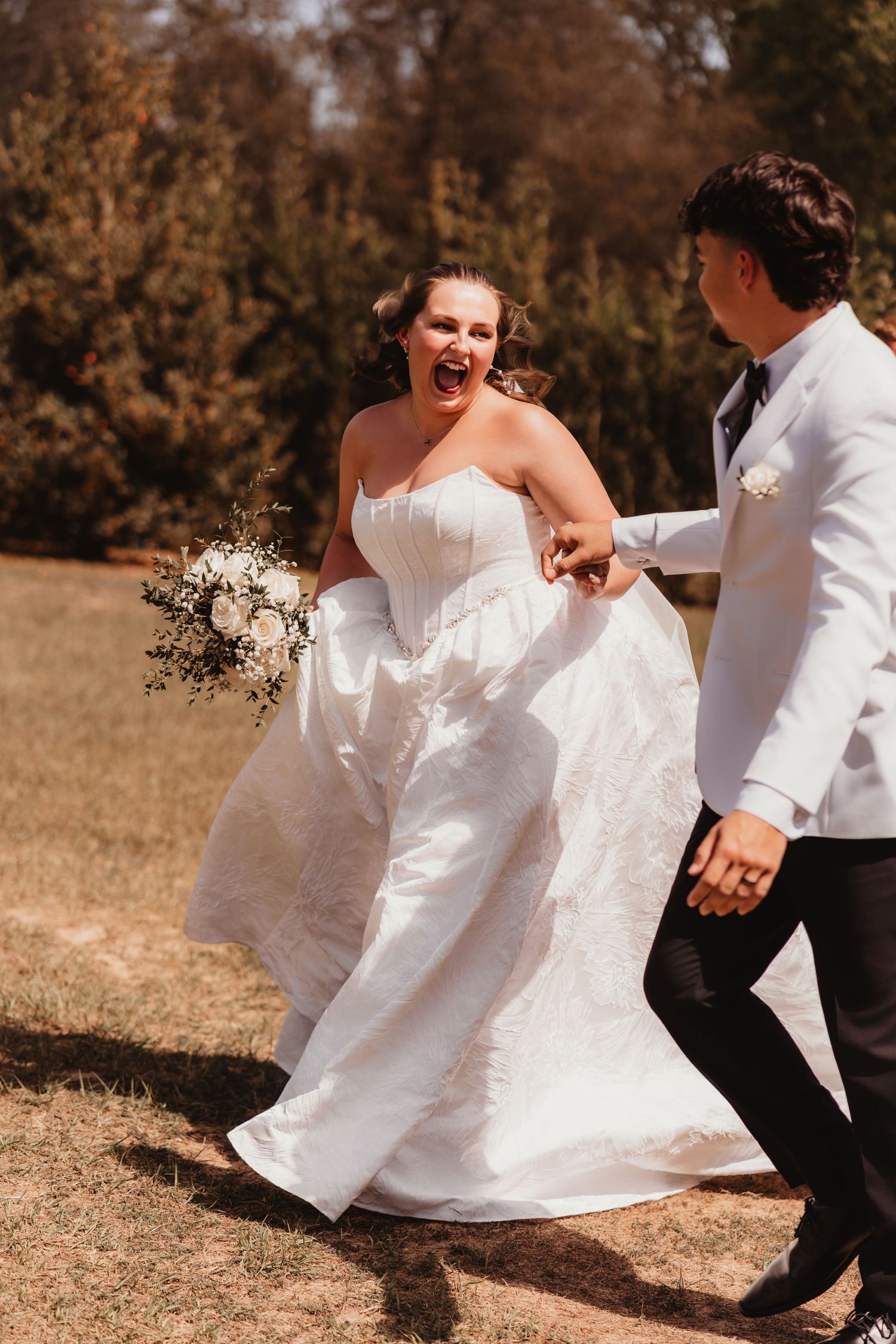 Excited bride in strapless white wedding gown laughing while running with groom in white tuxedo outdoors.