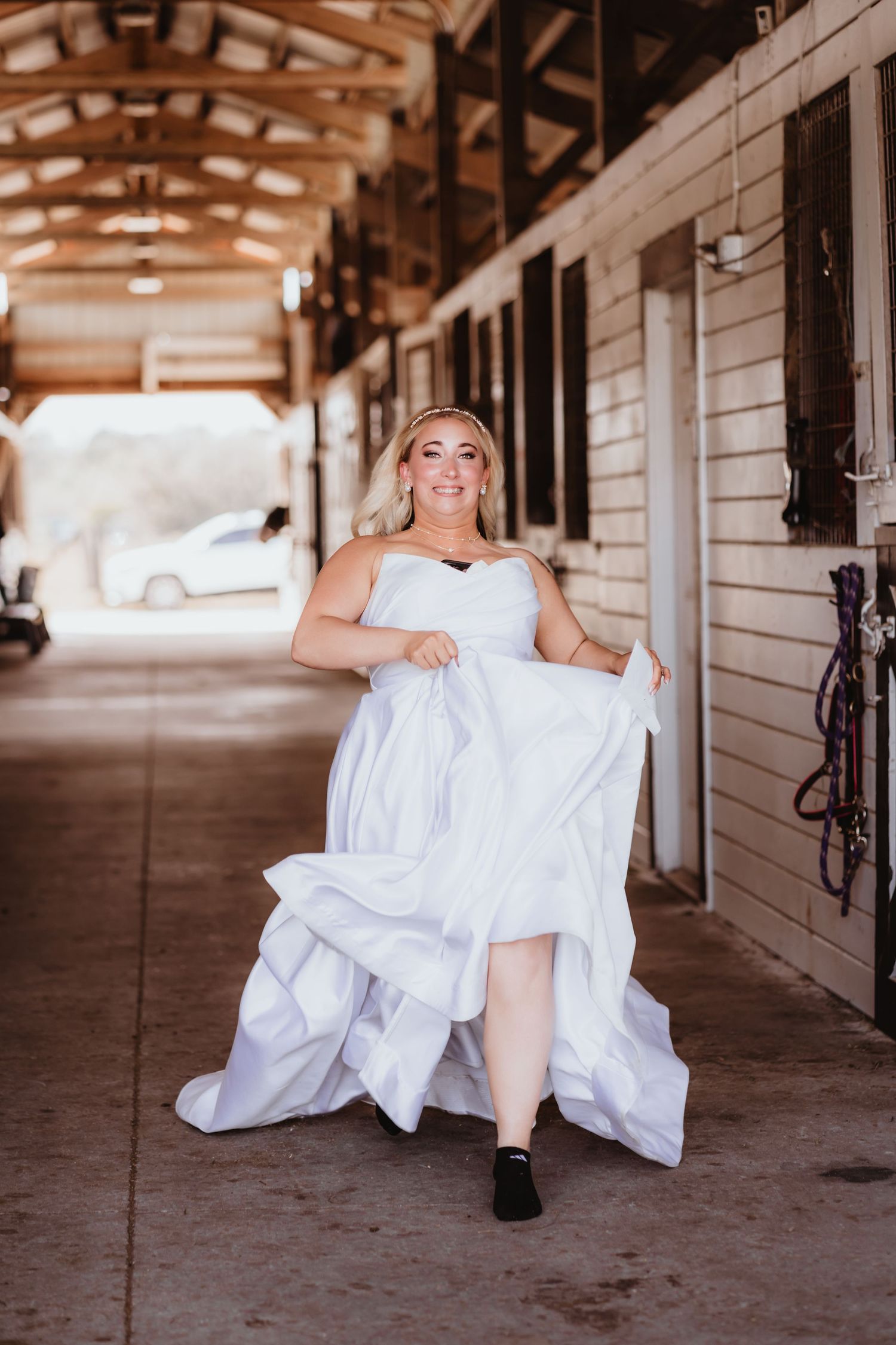 Joyful bride in strapless white ball gown lifts her skirt while walking through a rustic wooden barn aisle.