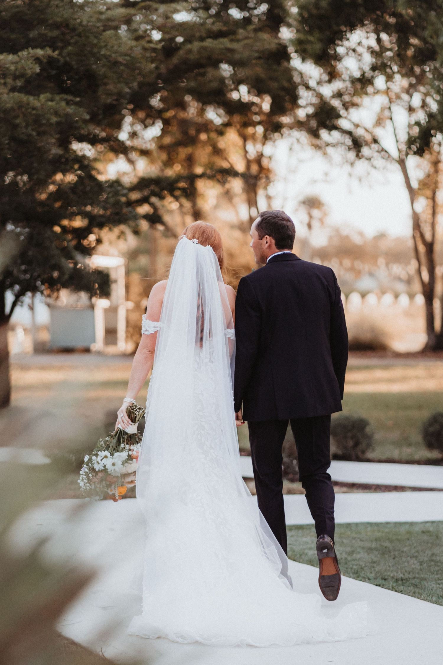 Bride and groom walk hand in hand along a garden path, her lace veil trailing behind her.
