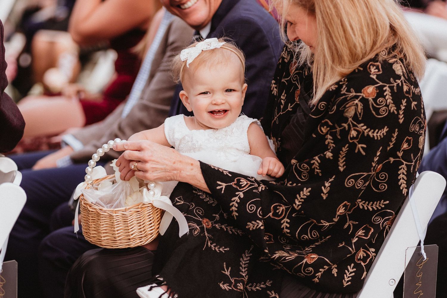 Adorable smiling baby flower girl in white dress holding a wicker basket at a wedding.