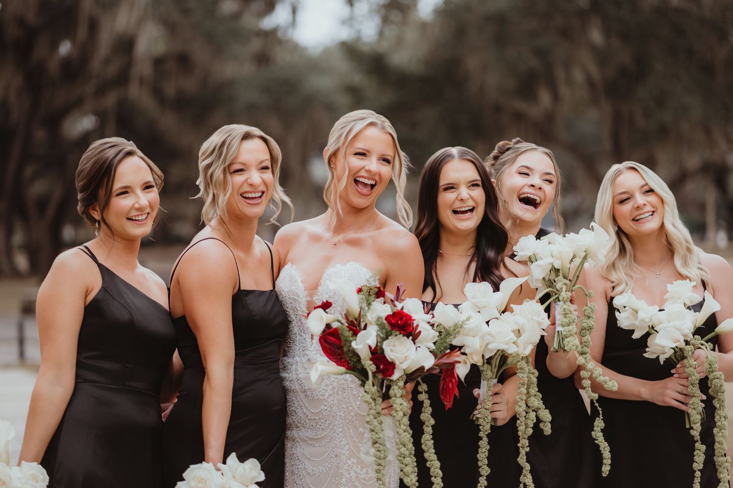 Laughing bride and bridesmaids in black dresses joyfully pose together holding white and red floral bouquets outdoors.